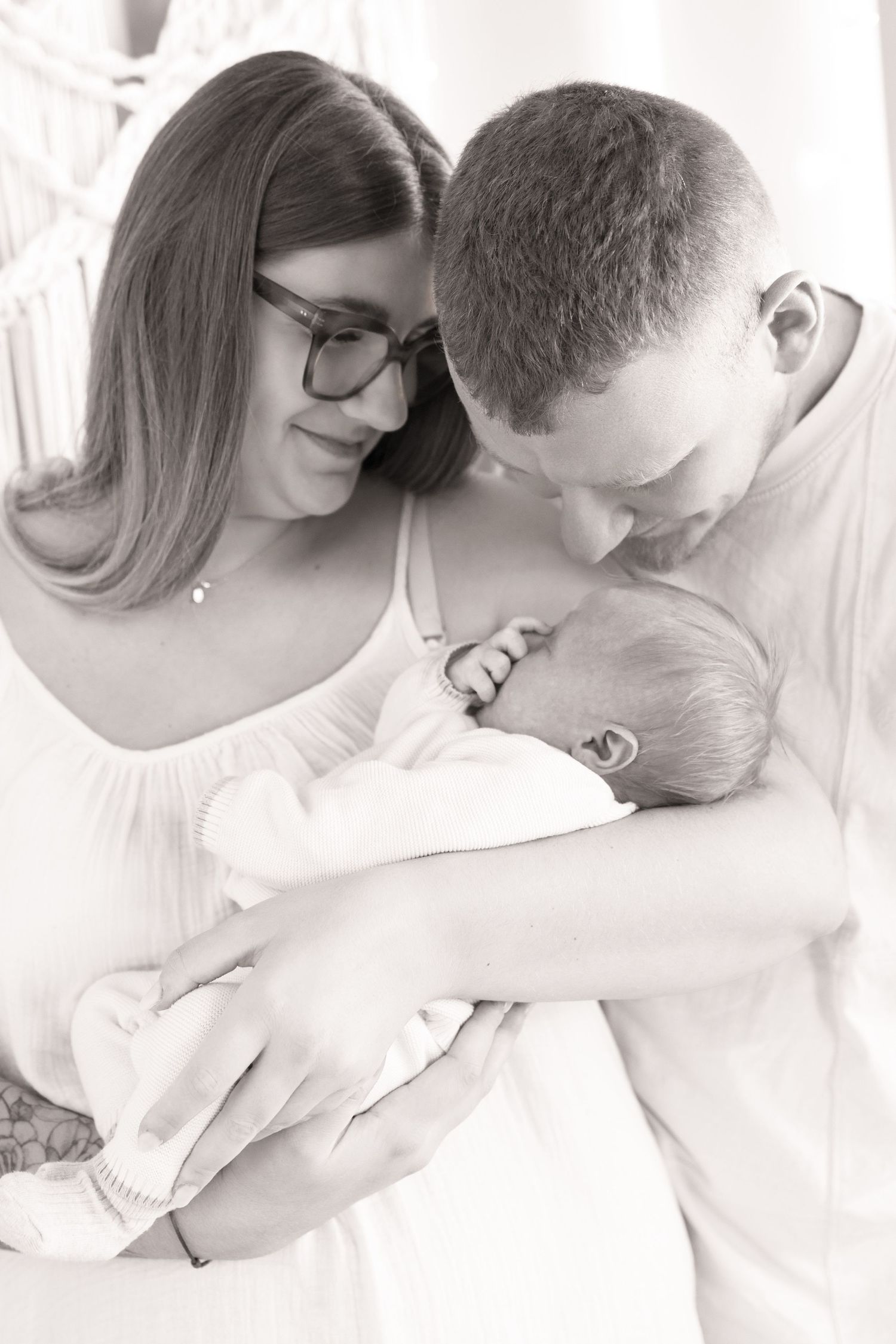 Black and white photo of new parents lovingly cradling their newborn baby together in a tender family portrait.