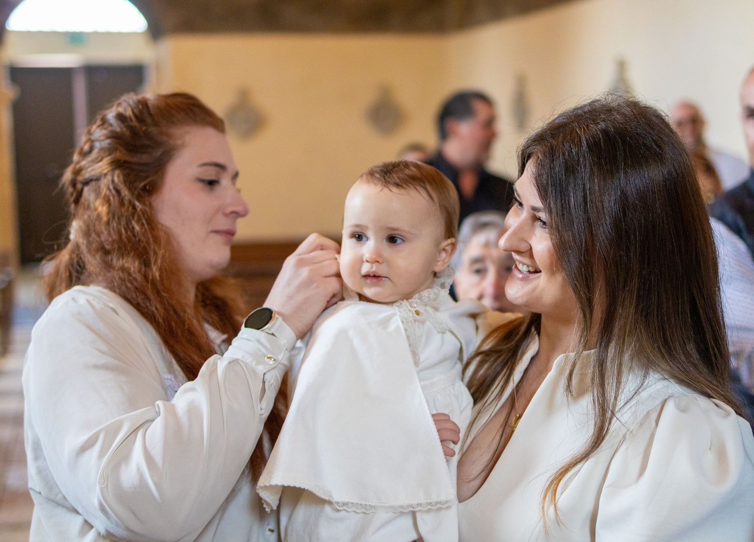 Two women in white dresses holding a baby in a christening gown inside a church.