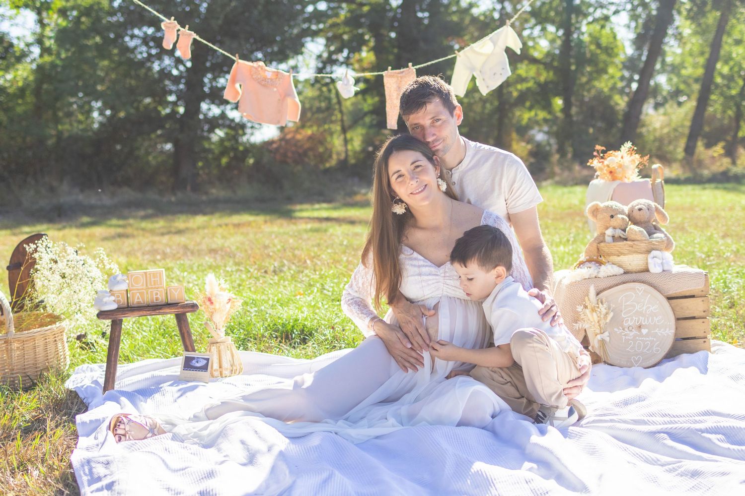 Family maternity photo shoot in a sunny park with parents and toddler on a white blanket with baby decorations.