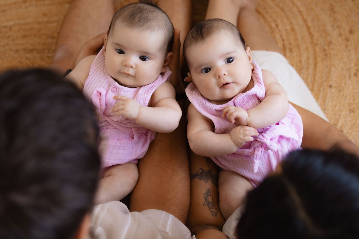 Two identical twin babies in pink onesies lying on an adult's lap, looking up curiously at the camera.
