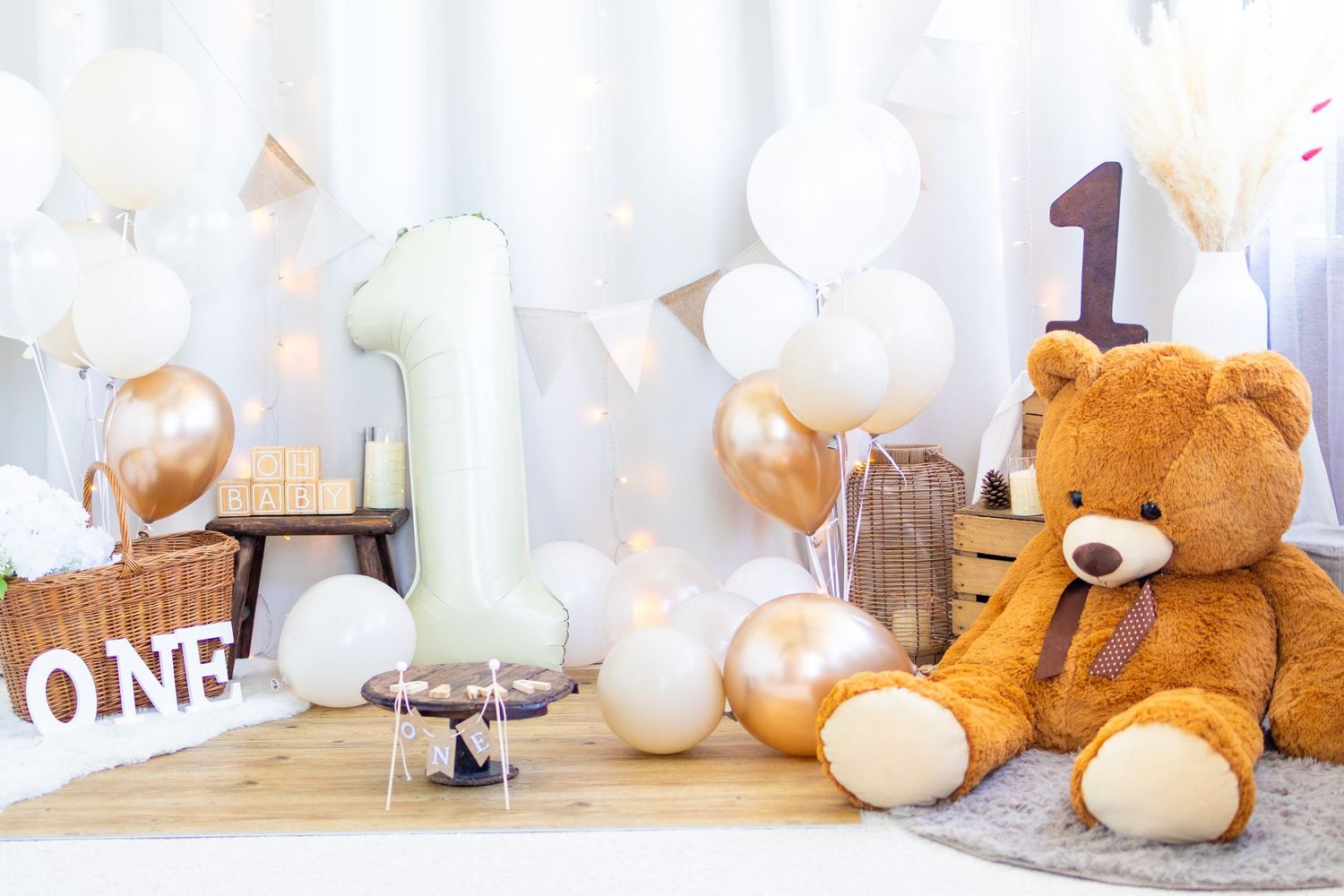 Teddy bear first birthday party setup with number one balloons, white and gold balloons, and ONE sign decoration.