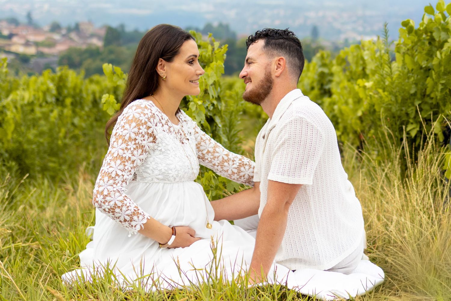 Pregnant woman in white lace dress sits with partner in green vineyard during maternity photo shoot.