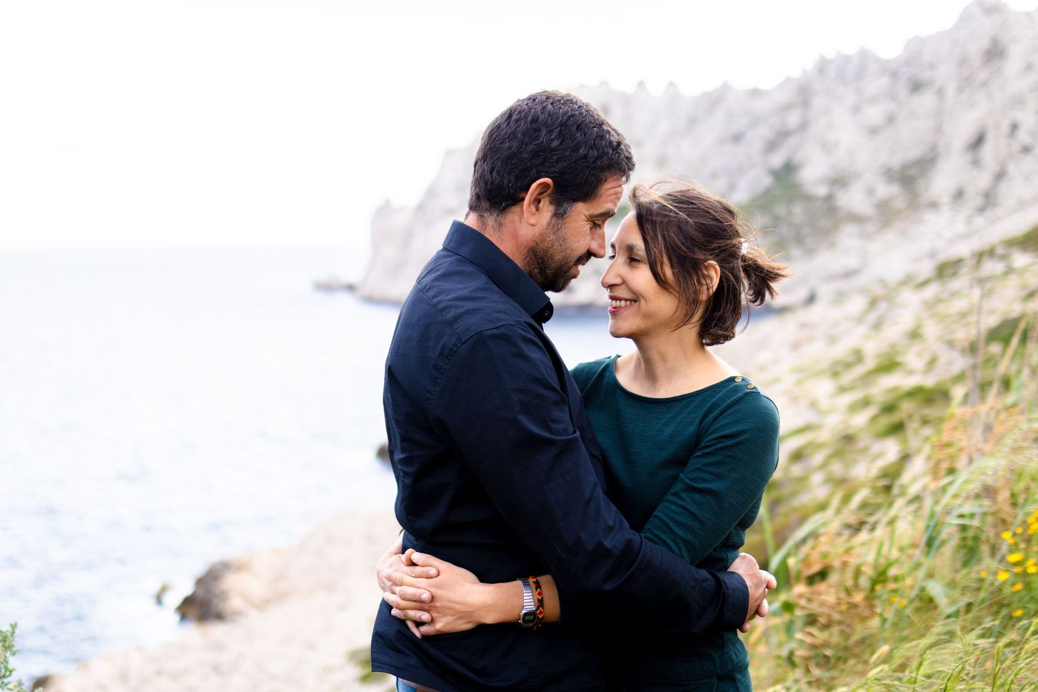 Couple embracing and smiling on a coastal cliff with ocean views on a cloudy day.
