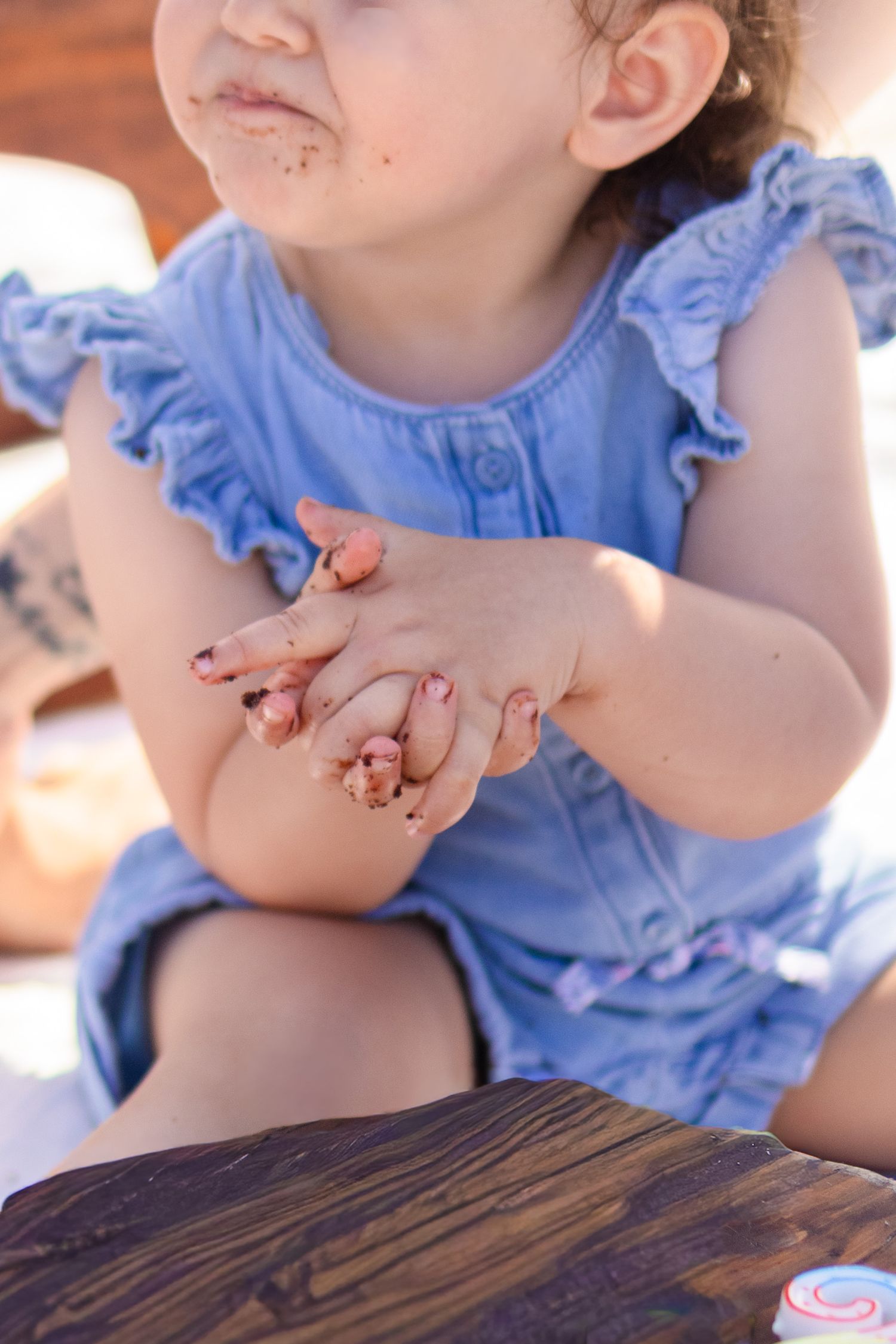 Toddler girl in blue ruffle romper with messy chocolate cake on her face and hands at birthday party.