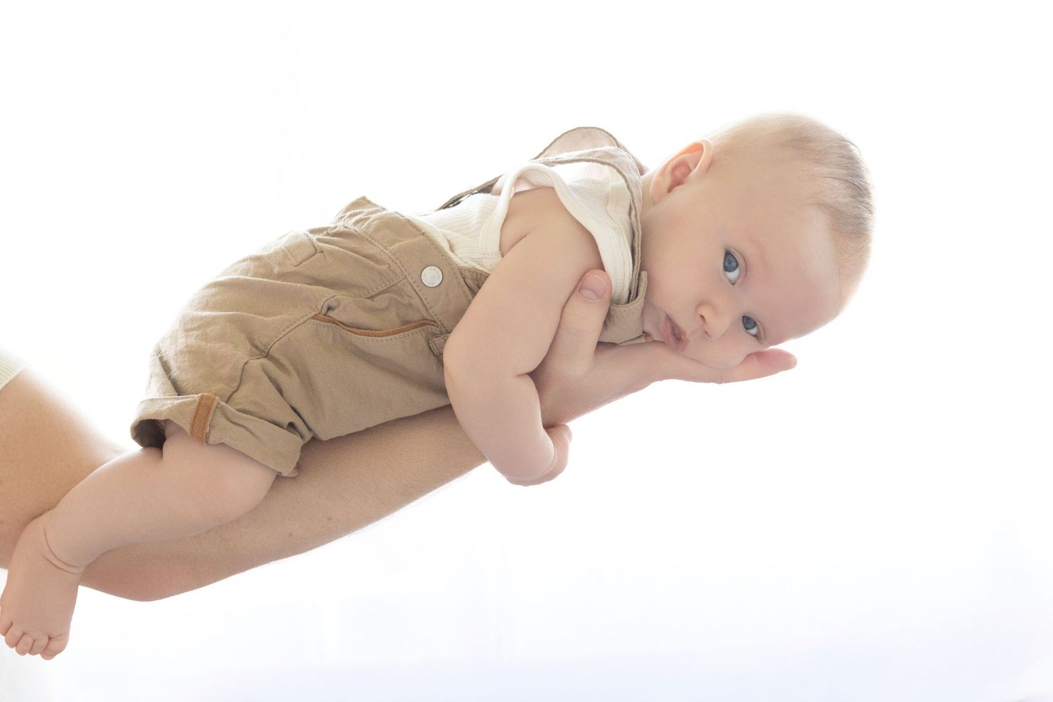 Adorable baby in tan romper being held horizontally by adult hands against white background.