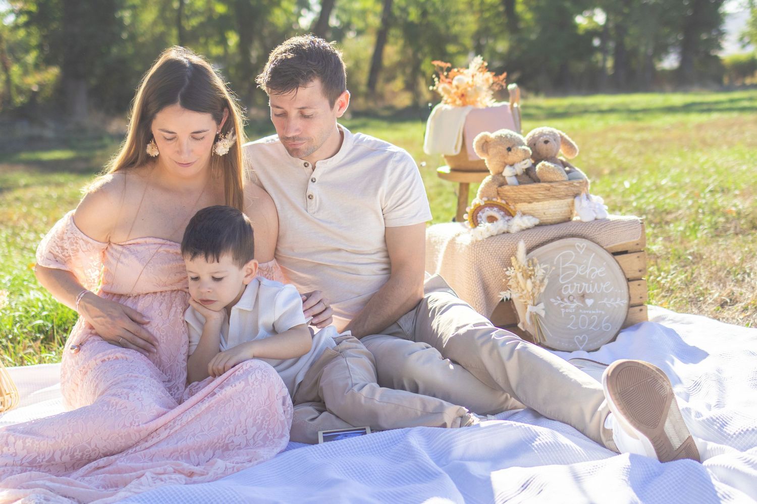 Pregnant woman and man sit on blanket outdoors with young boy during sunny family maternity photo session.