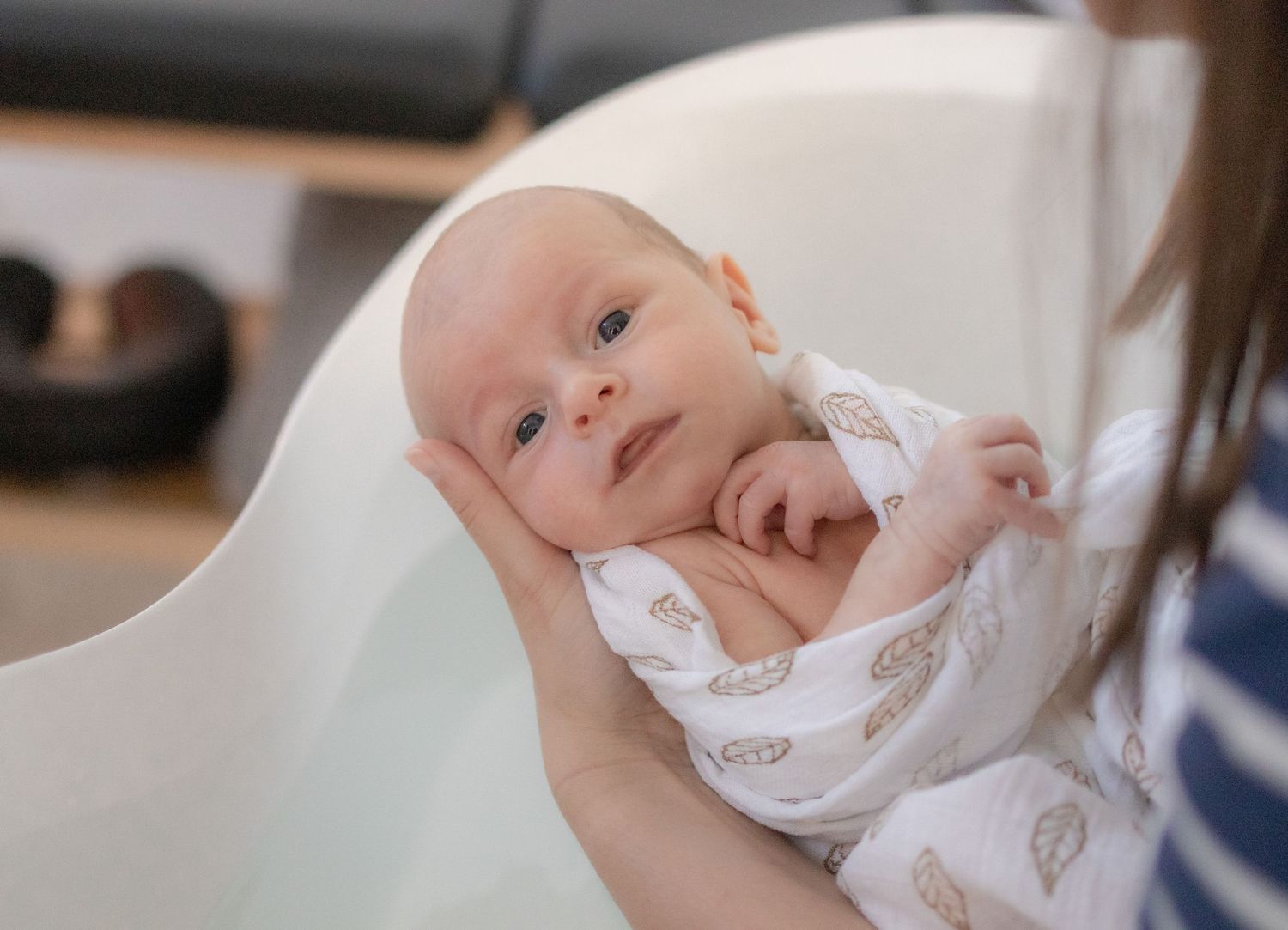 Newborn baby wrapped in white muslin swaddle blanket being held by mother in bright modern nursery.