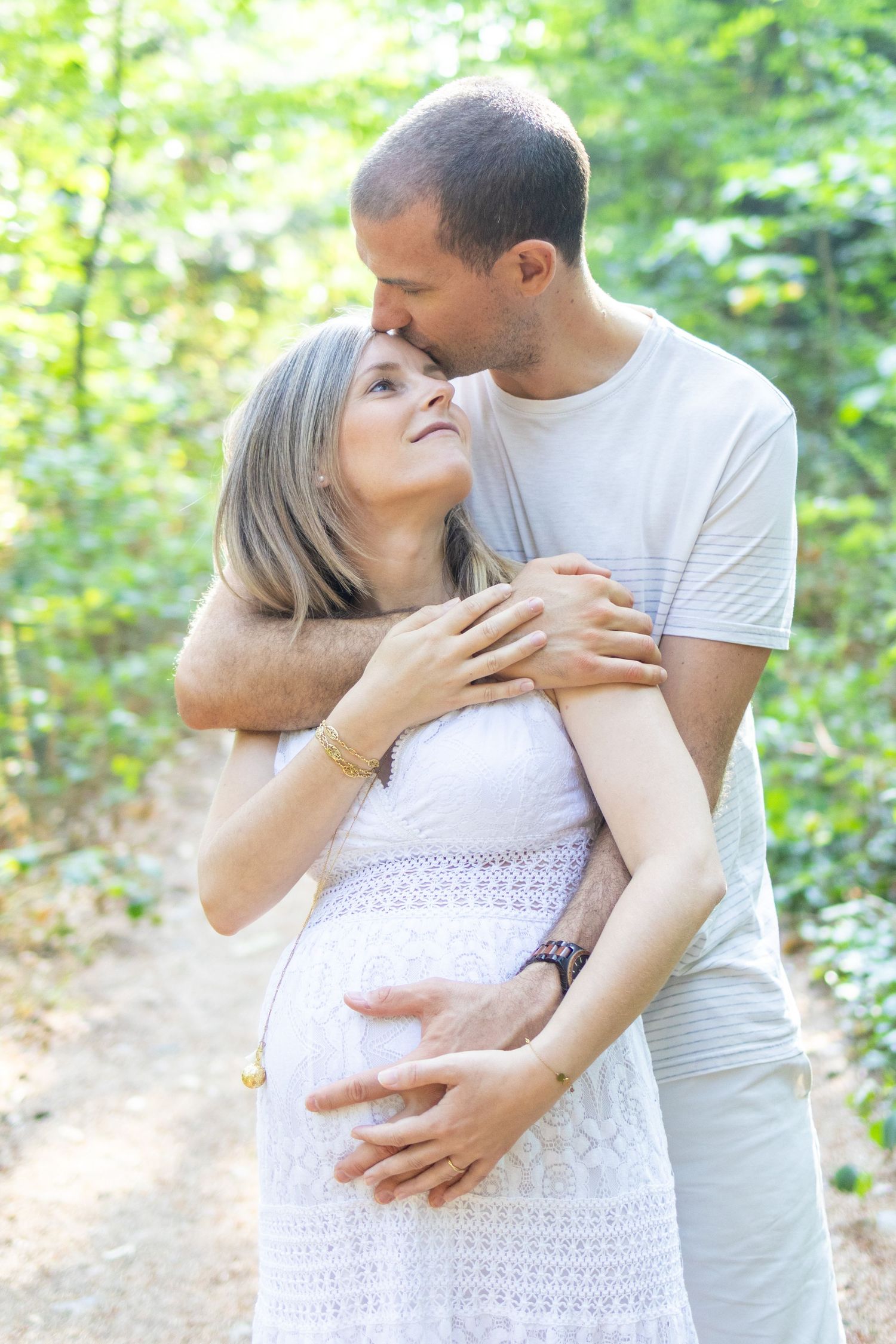 A man tenderly embraces a pregnant woman in a white dress outdoors, kissing her forehead on a sunny forest path.