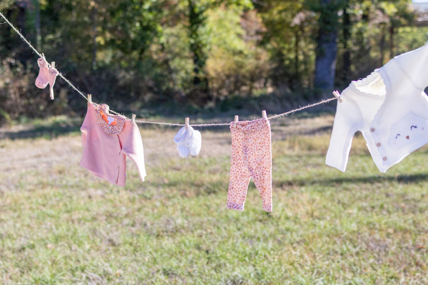 Baby clothes hanging on a clothesline outdoors in a sunny green field, including pink onesie, floral pants, and white shirt.