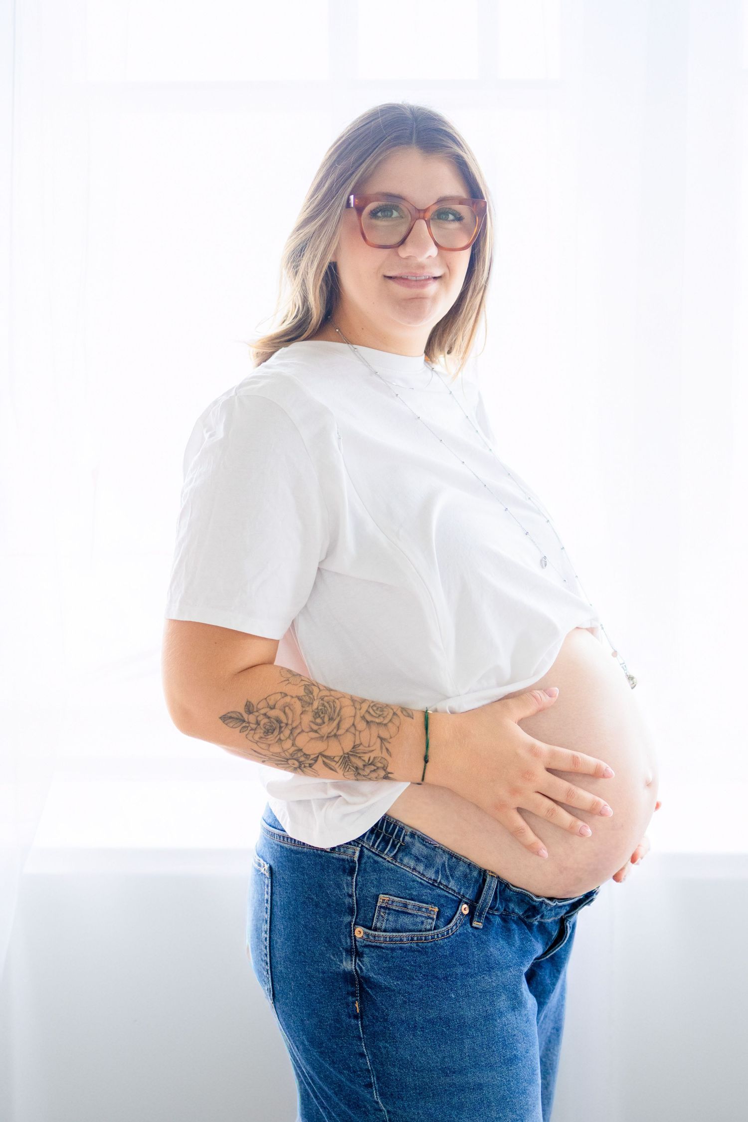 Smiling pregnant woman in white shirt and jeans, holding baby bump, wearing glasses, with floral tattoo on arm.