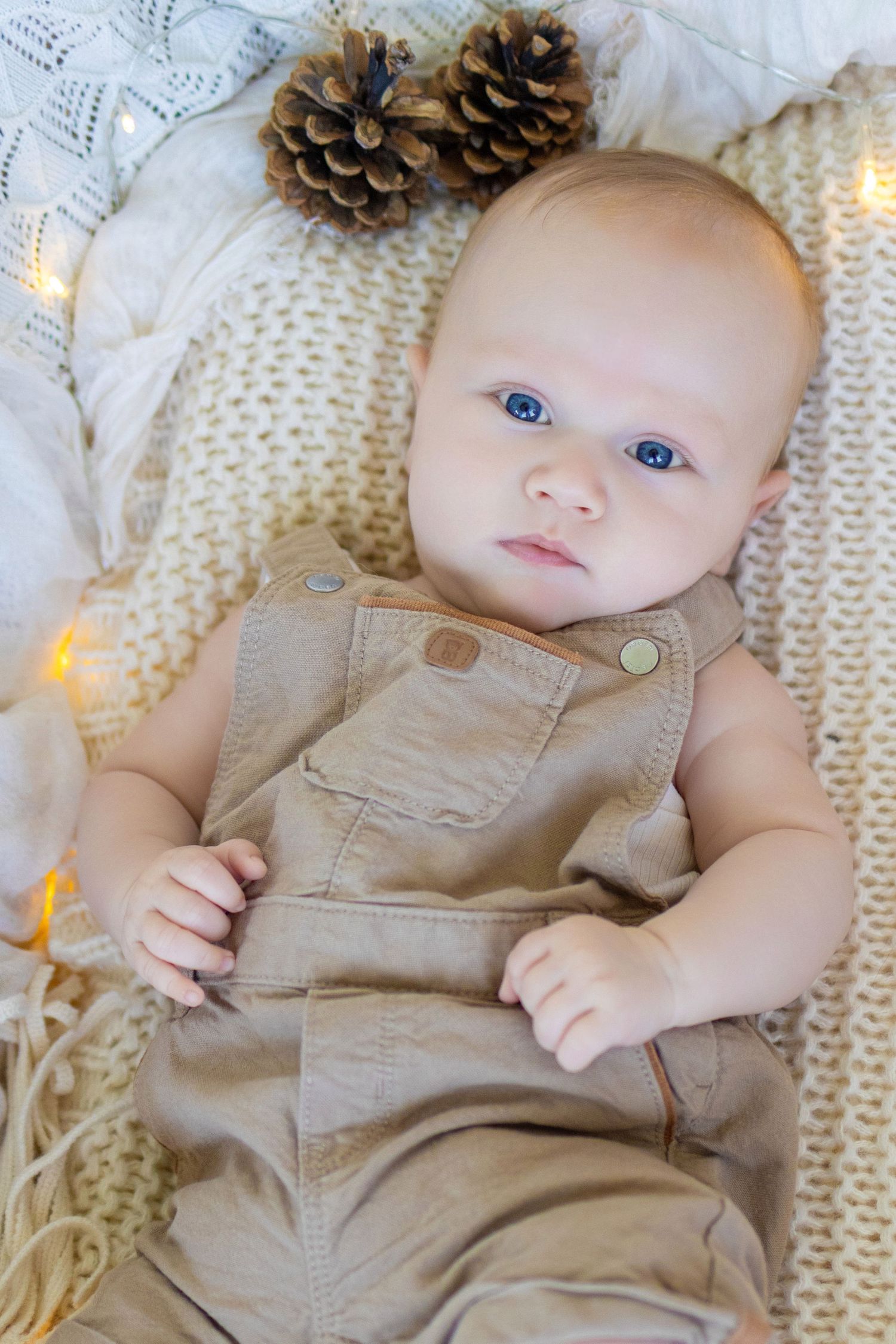 Blue-eyed baby in tan overalls lying on knit blanket with pine cones and warm fairy lights.