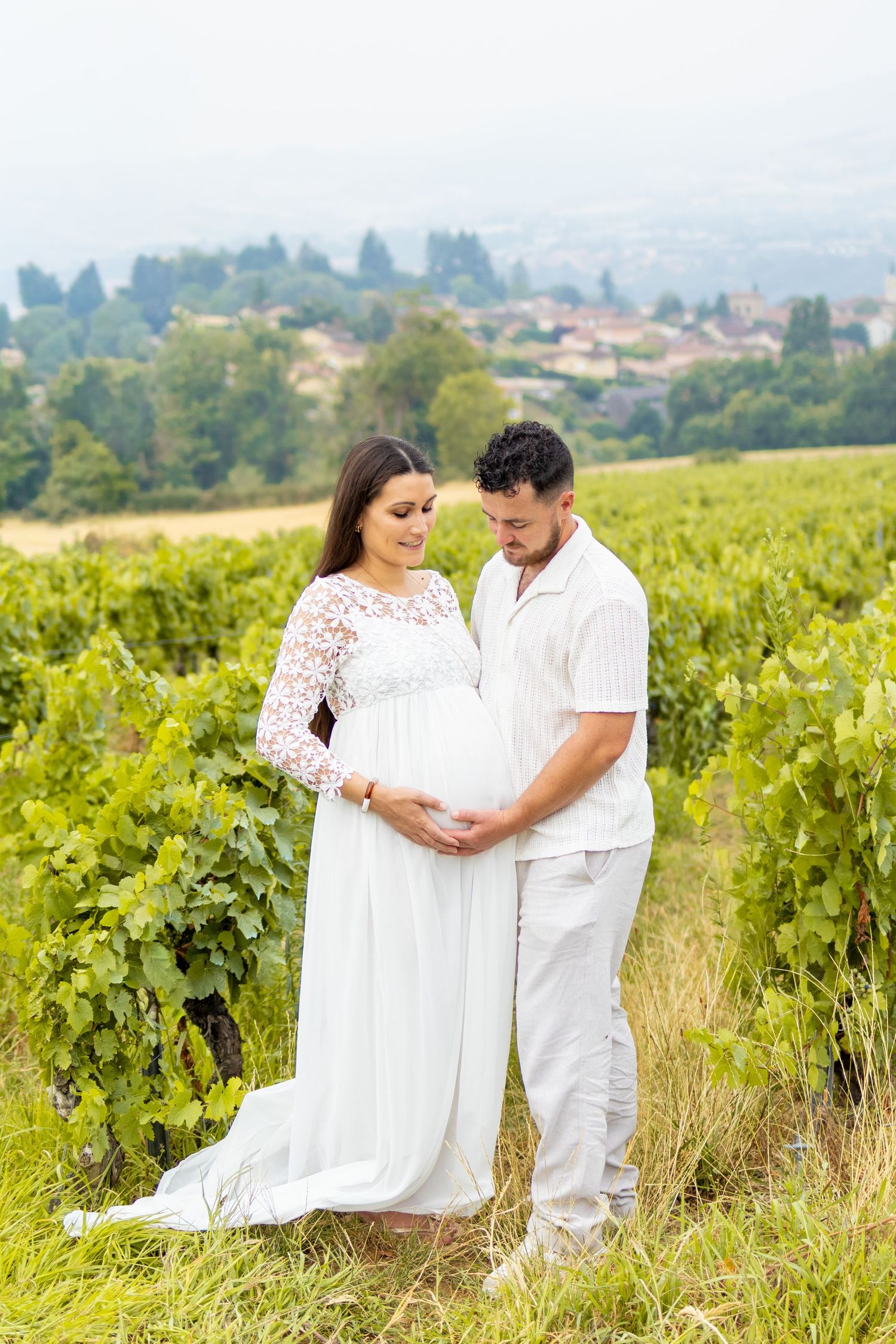 Couple in white outfits tenderly cradling pregnant belly in lush vineyard with scenic village backdrop.