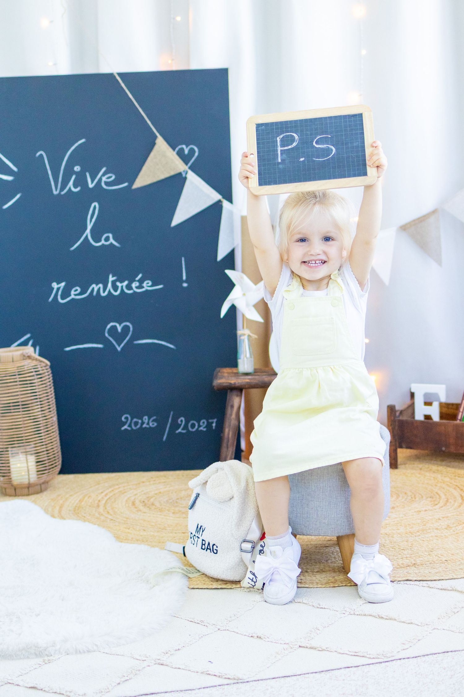 Smiling blonde toddler girl in yellow dress holds up a small chalkboard with PS written on it, with Vive la rentree backdrop.
