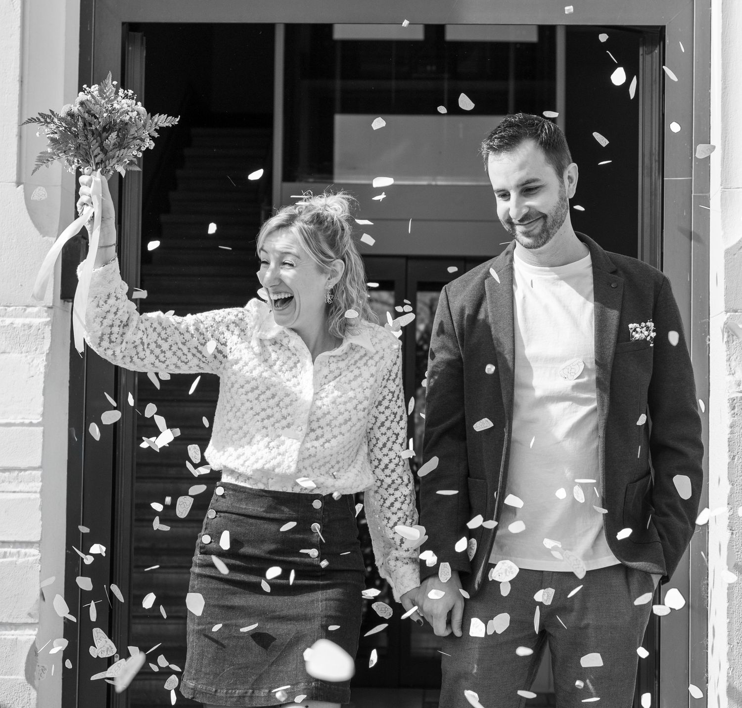 Black and white photo of a joyful couple celebrating their wedding exit with confetti, bride holding a bouquet.