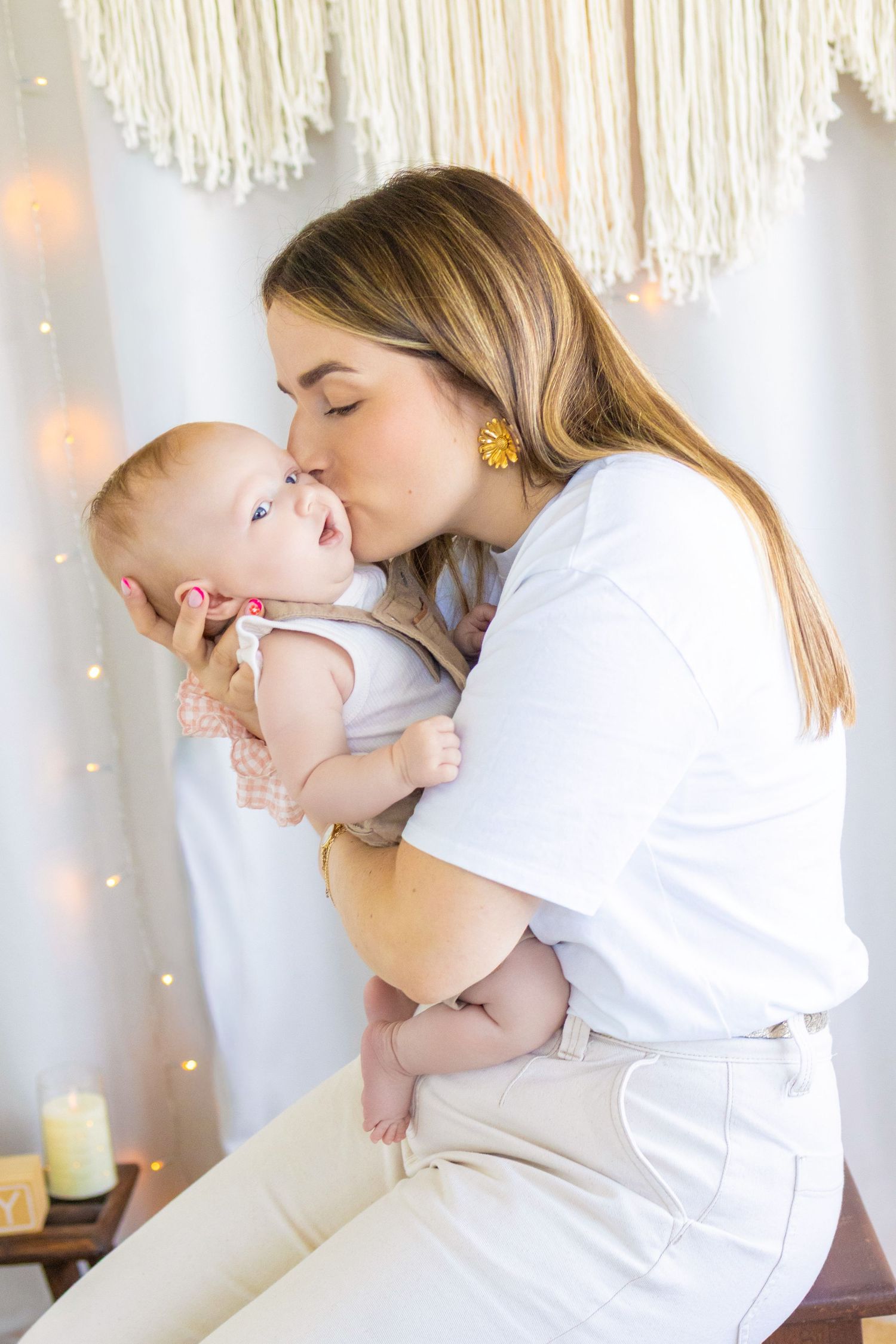 A young mother tenderly kisses her infant baby while holding them in a warmly lit, bohemian-styled room.