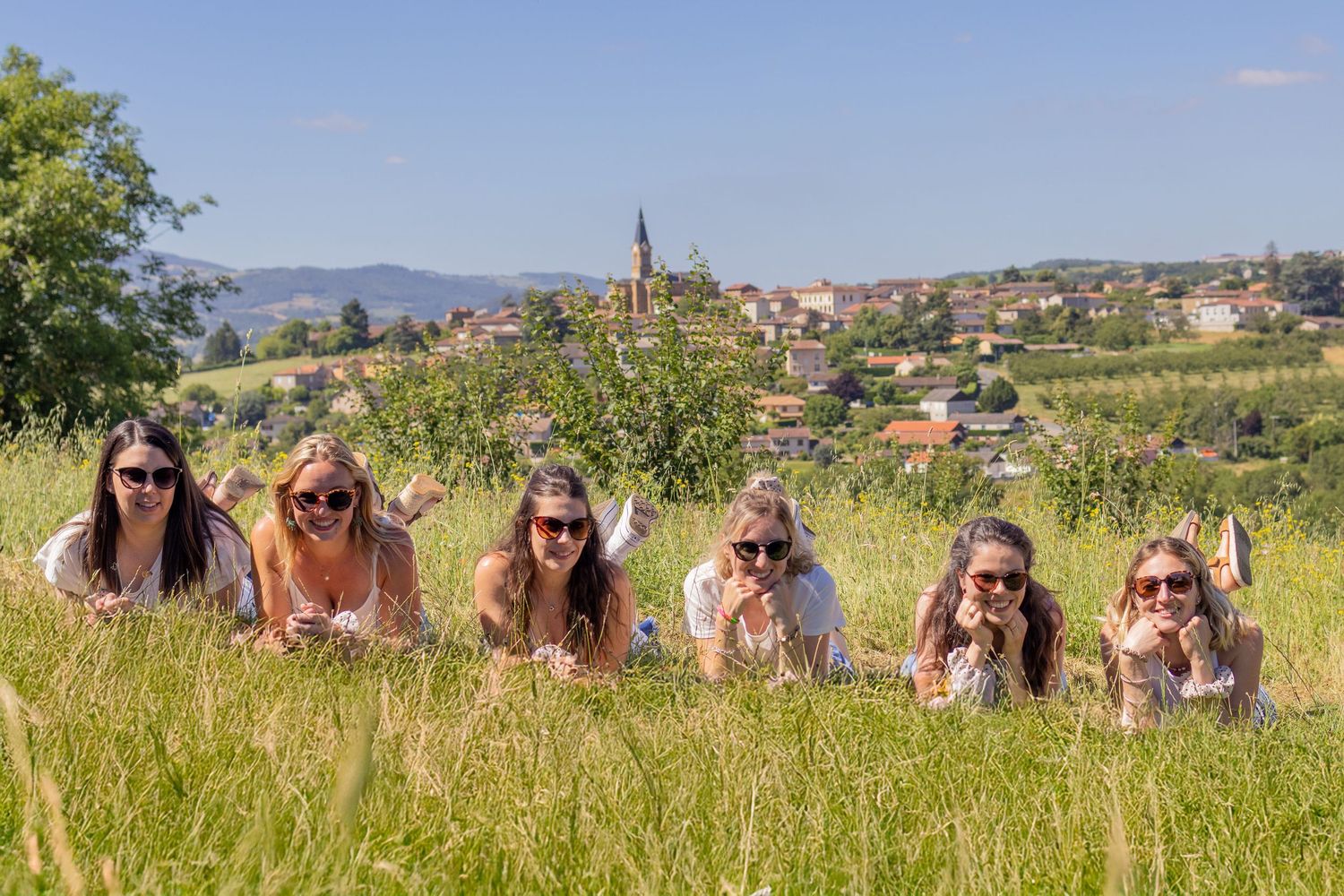 Six smiling women lying in a green meadow with a charming European village and church steeple in the sunny background.