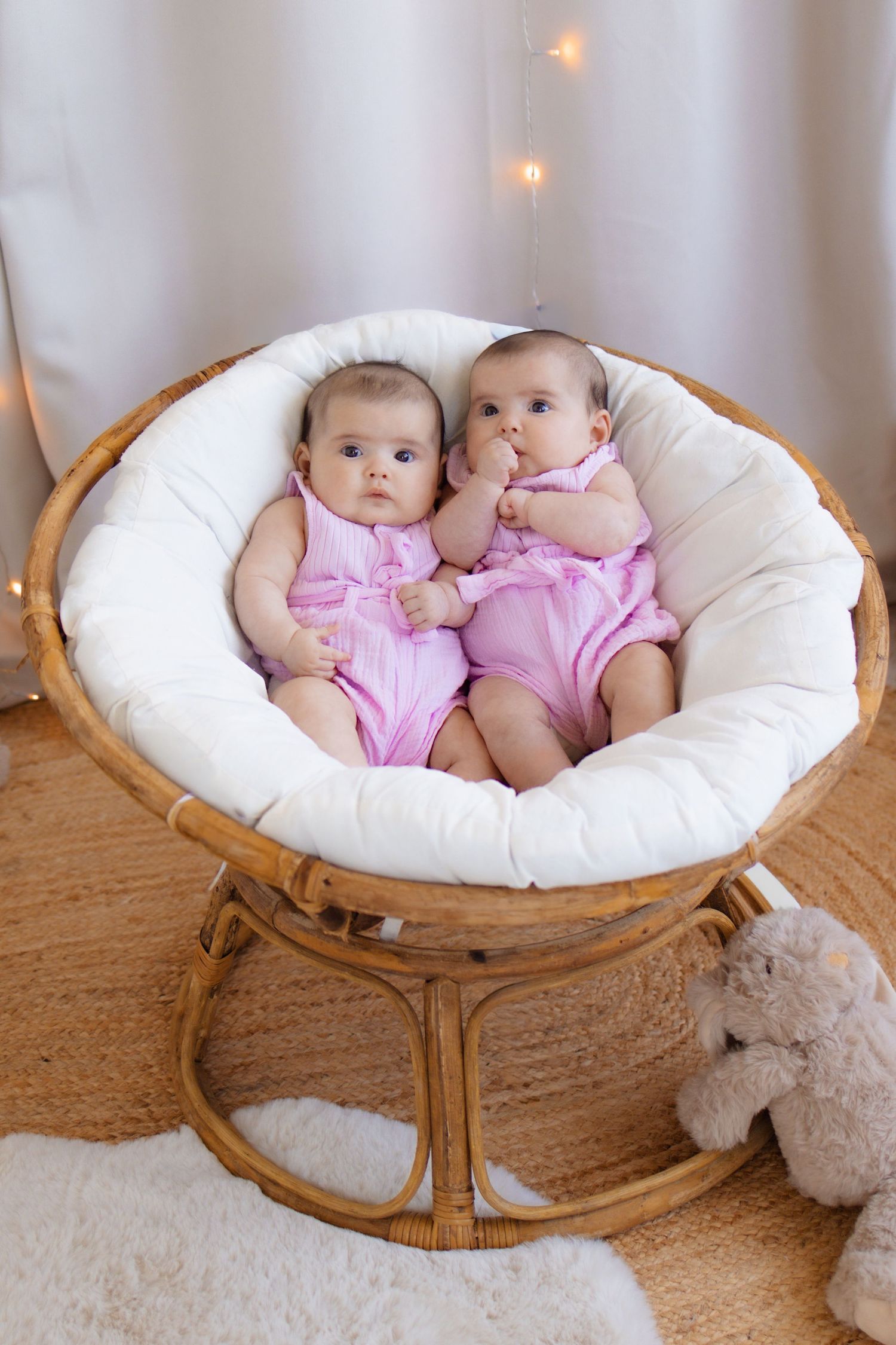 Two adorable twin babies in matching pink rompers sitting together in a round rattan papasan chair with white cushion.