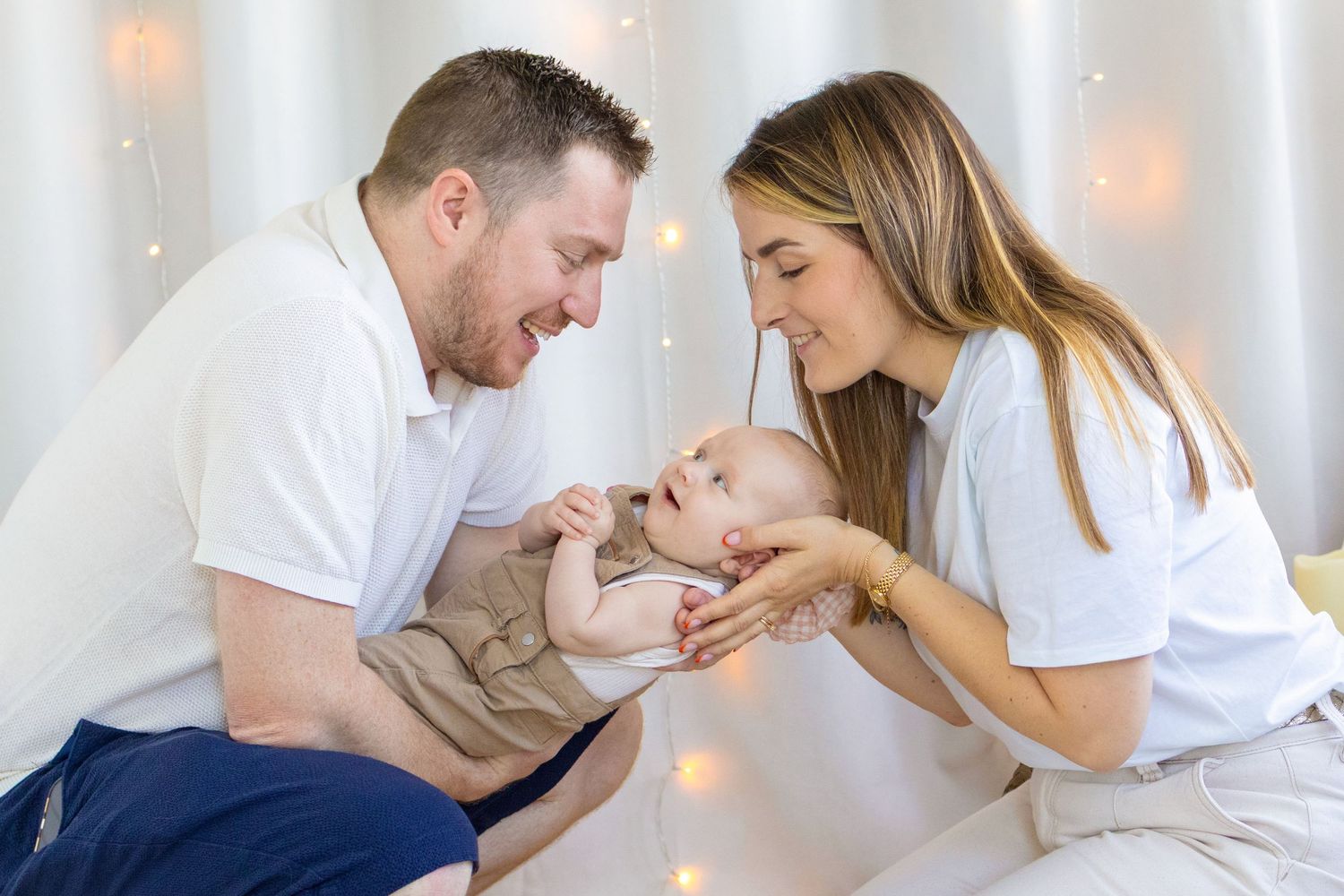 Happy parents in white shirts smiling and playing with their newborn baby against a soft bokeh light background.