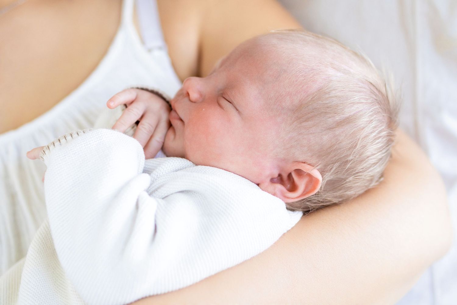 Newborn baby sleeping peacefully in mother's arms, dressed in white clothing.
