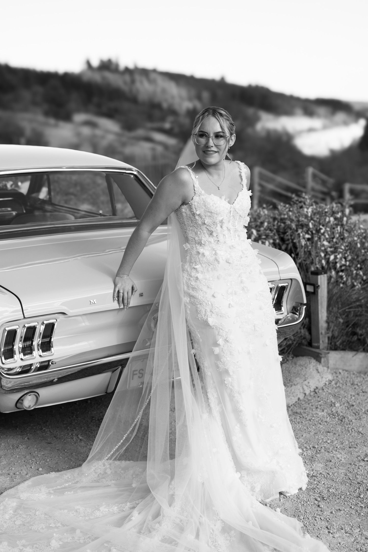 Bride in floral wedding gown with tulle train poses beside vintage Ford Mustang in black and white outdoor photo.