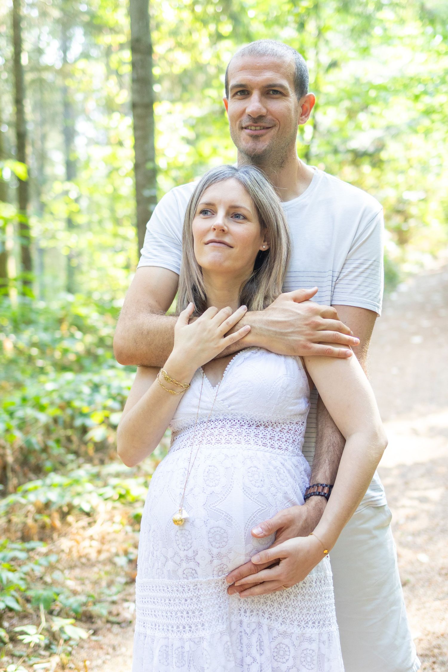 A man embraces a pregnant woman in a white dress outdoors in a sunlit forest setting.