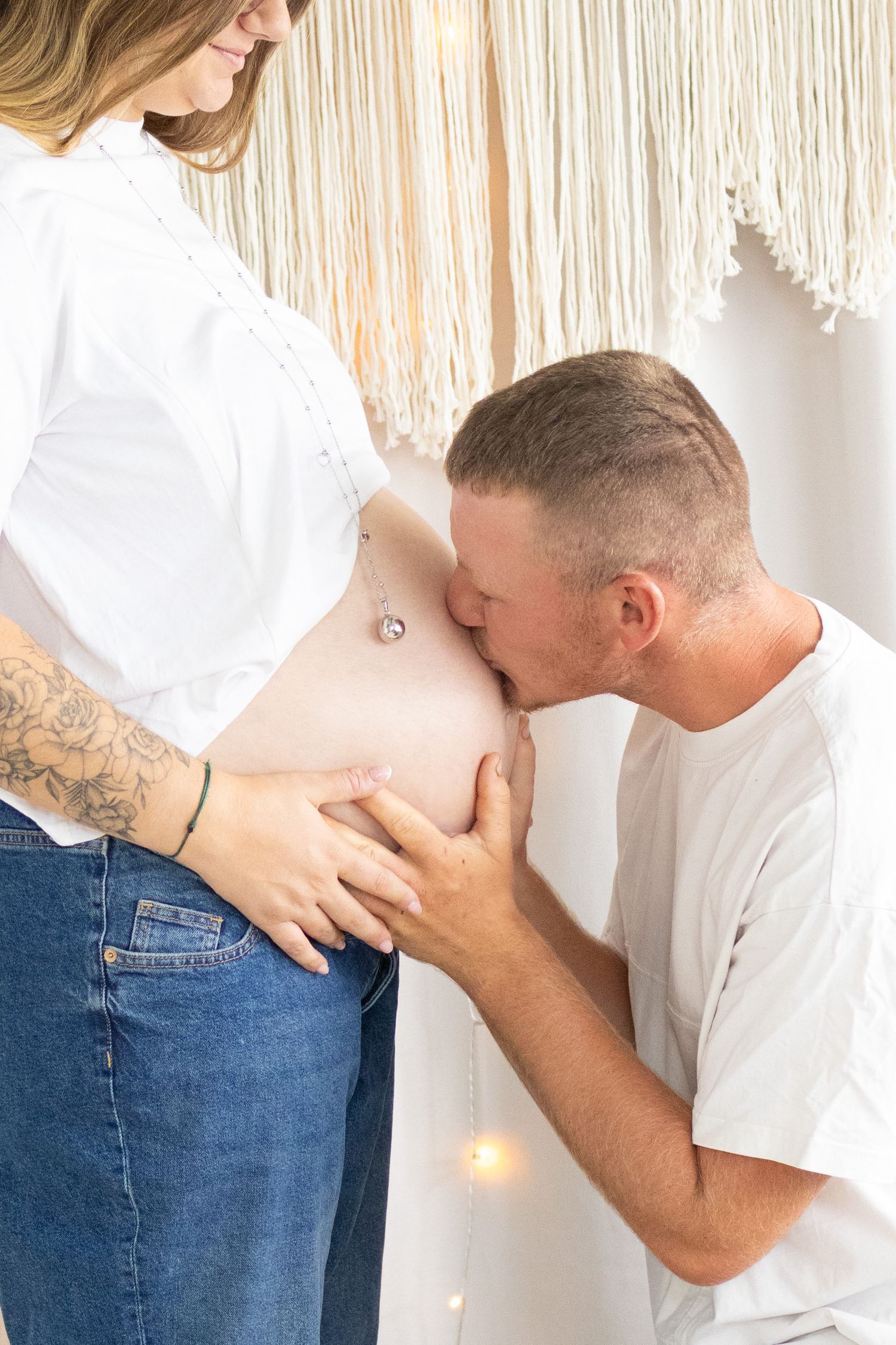 A man kisses his pregnant partner's baby bump while she smiles, both wearing white tops in a bohemian-styled room.