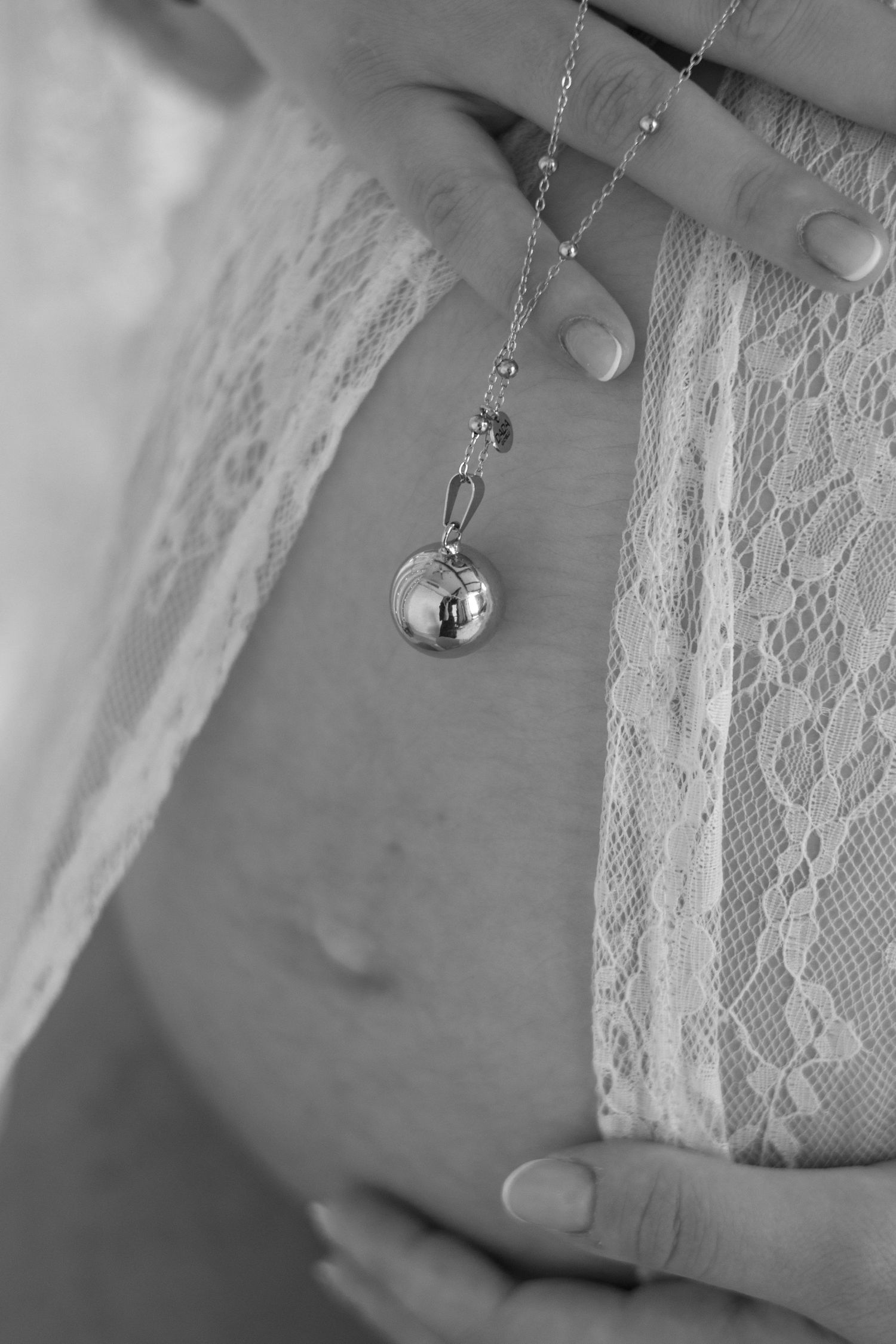 Black and white photo of pregnant woman wearing lace top, holding belly with a silver bola pregnancy necklace.