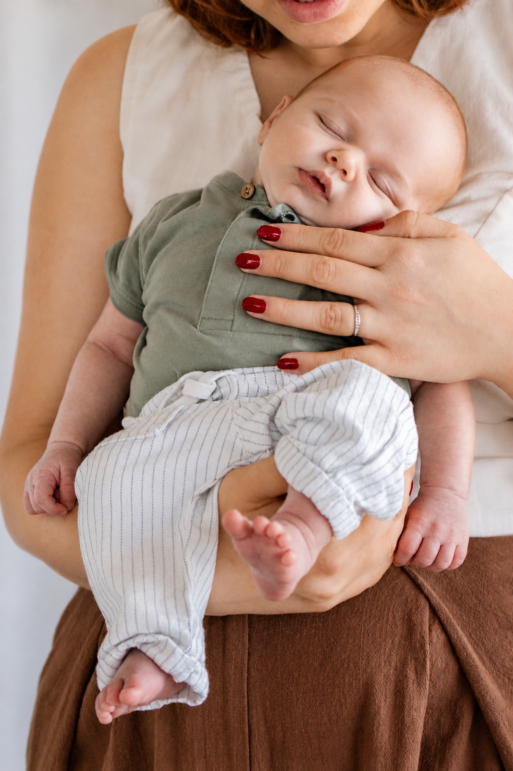 A mother with red nails gently holds her sleeping newborn baby dressed in a green onesie and striped pants.