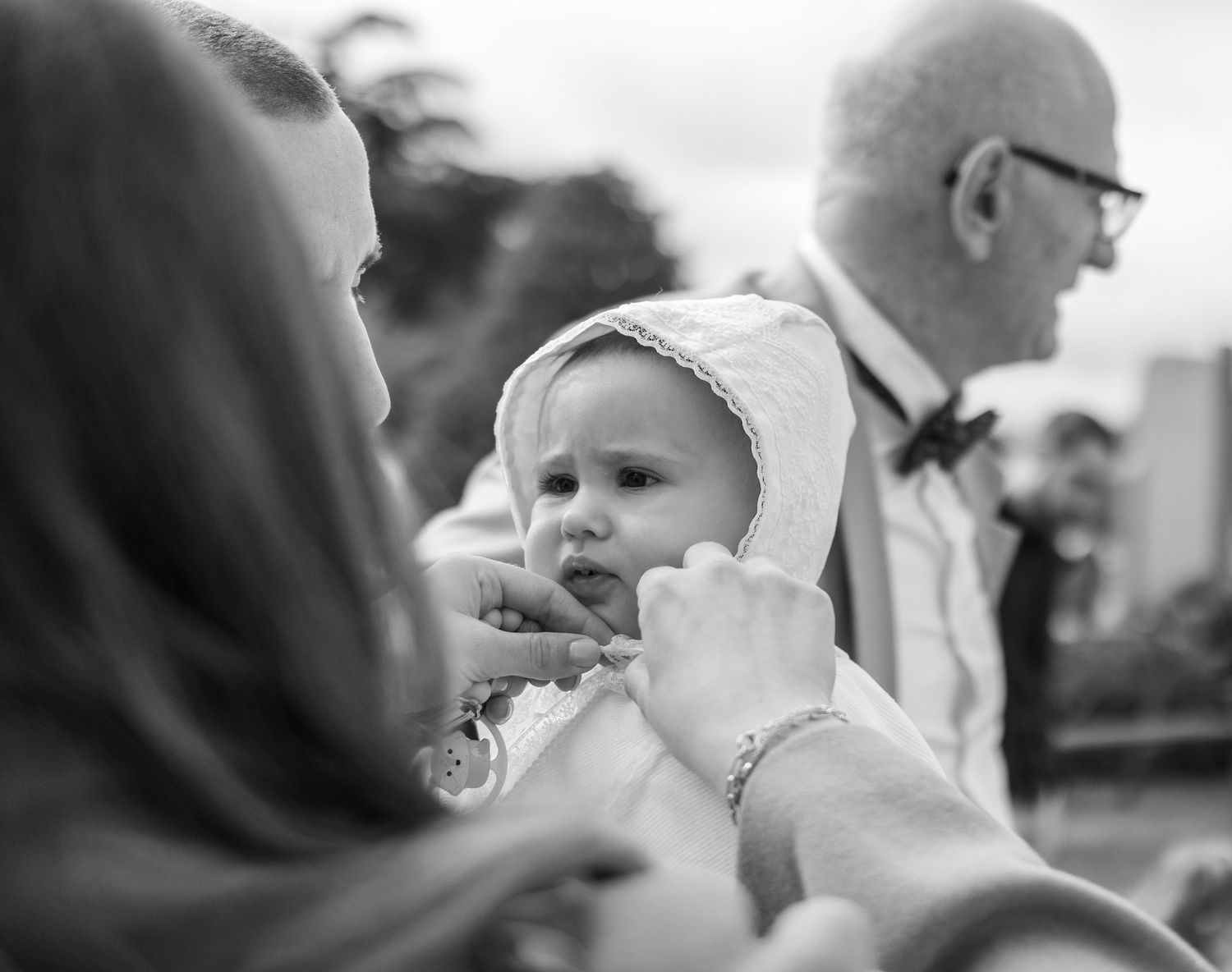 Black and white photo of a baby in a bonnet being held at a formal event with adults in the background.