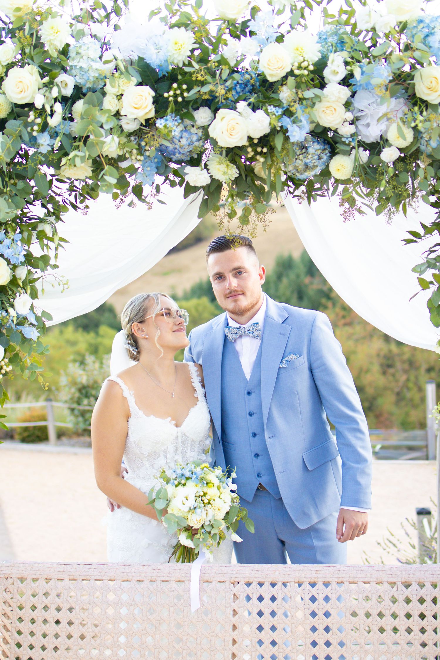 Bride in white lace gown and groom in light blue suit stand under floral arch with white and blue blooms.