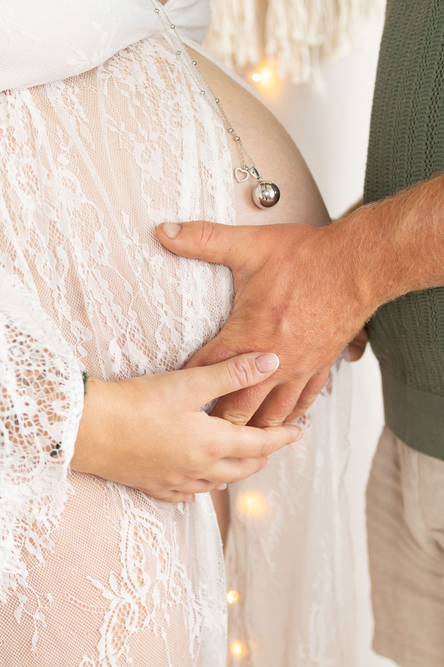 Pregnant woman in white lace dress with partner's hand gently resting on her baby bump, adorned with a delicate necklace.