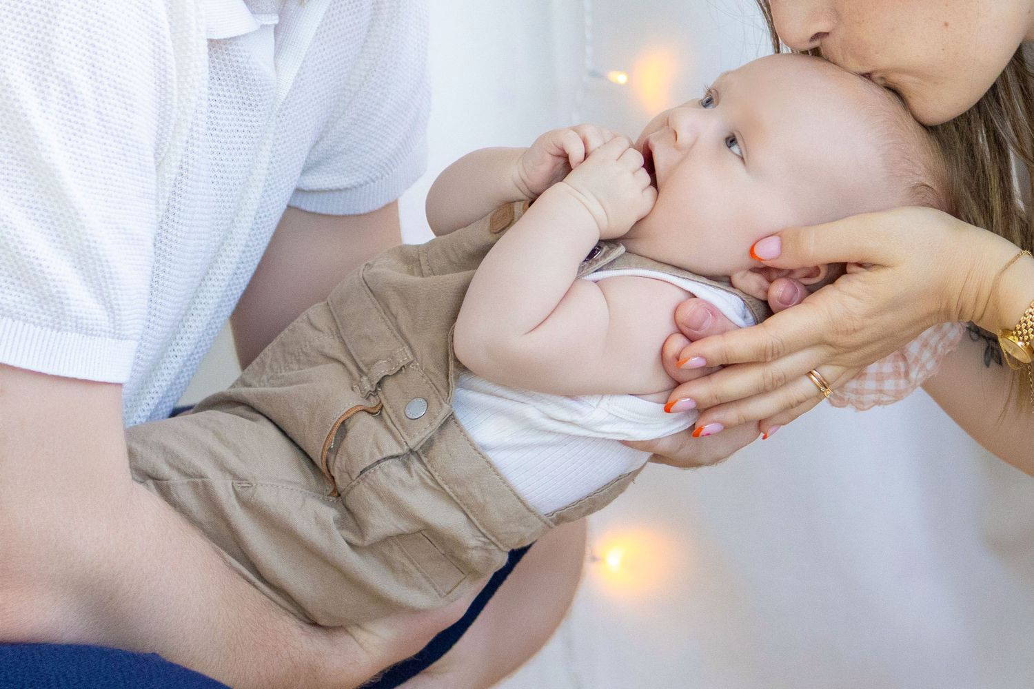 Parents lovingly kissing and holding their smiling baby boy dressed in white onesie and khaki shorts.