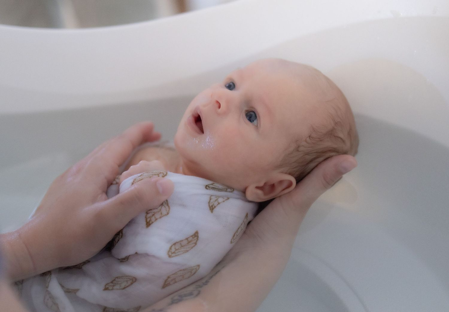 Newborn baby in a patterned wrap being gently bathed in a white tub, supported by adult hands.