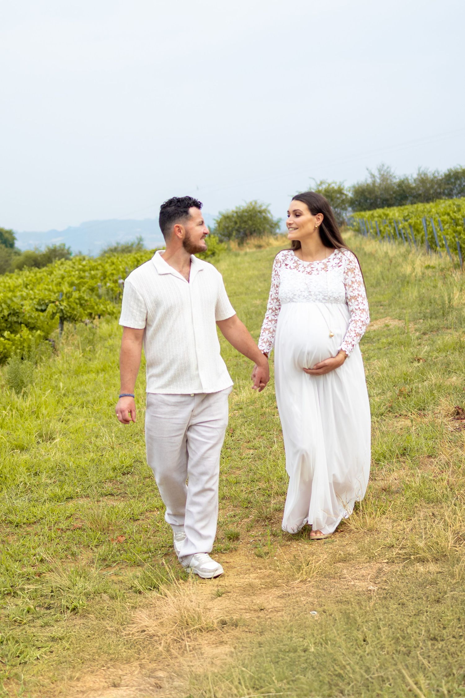Pregnant woman in white dress walks hand-in-hand with man in white outfit through lush green vineyard.