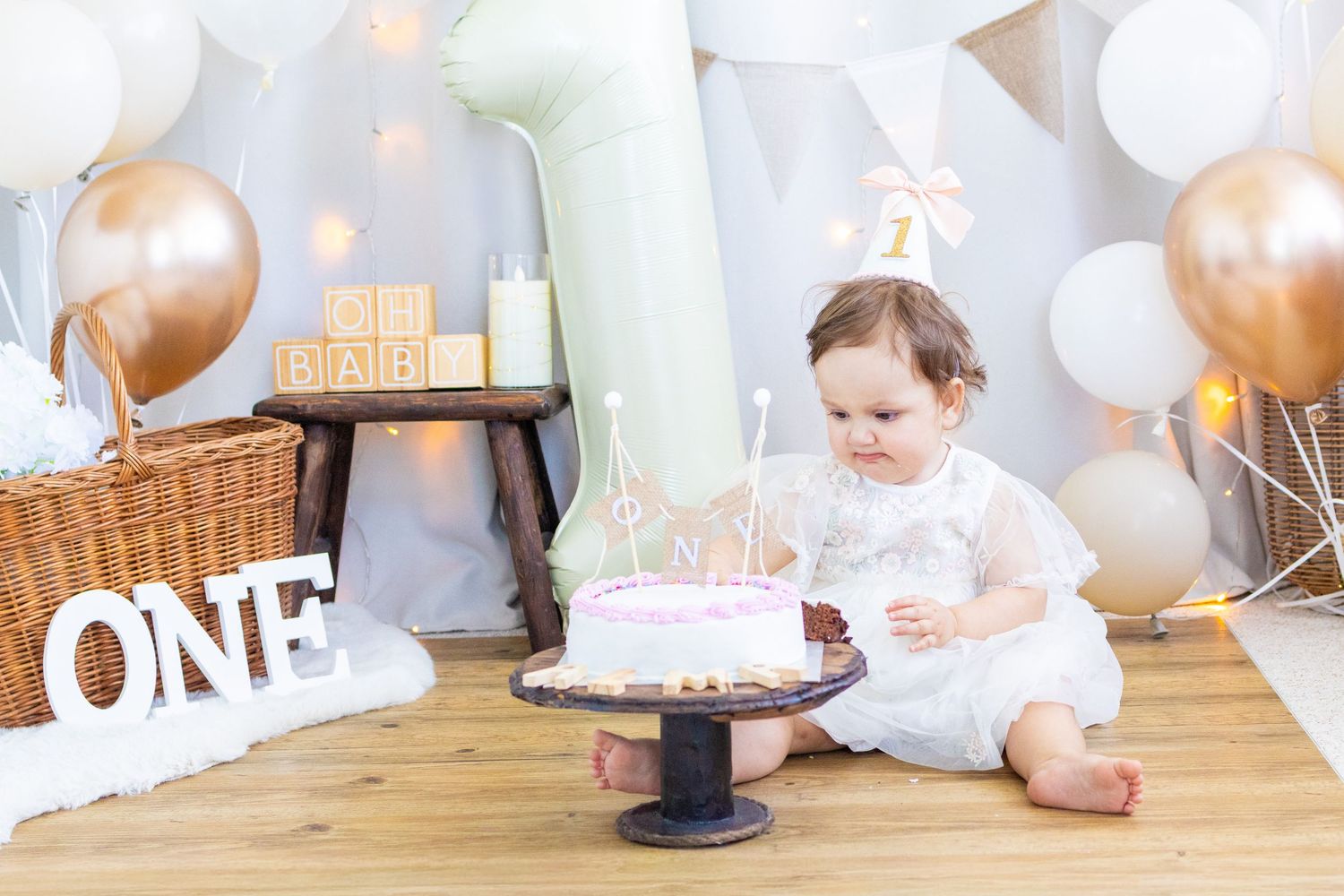 Baby girl celebrating her first birthday with a pink cake, balloons, and ONE sign in a decorated photo studio.