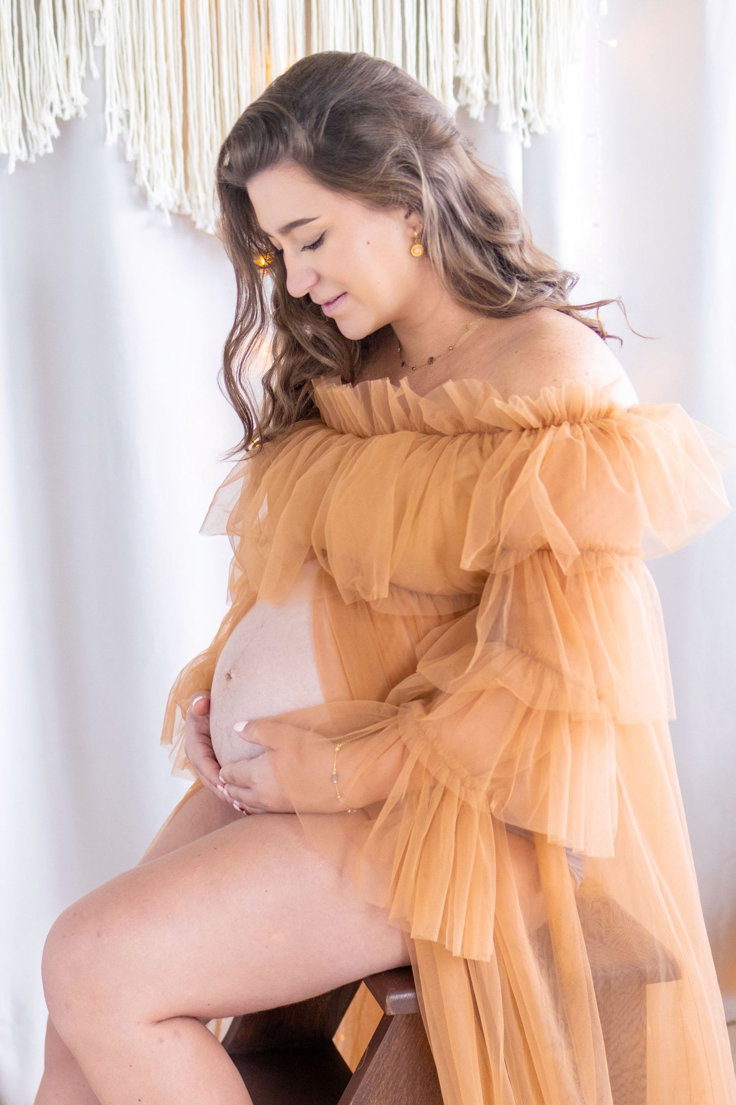 Pregnant woman in elegant orange tulle off-shoulder gown cradling baby bump, seated against white boho backdrop.