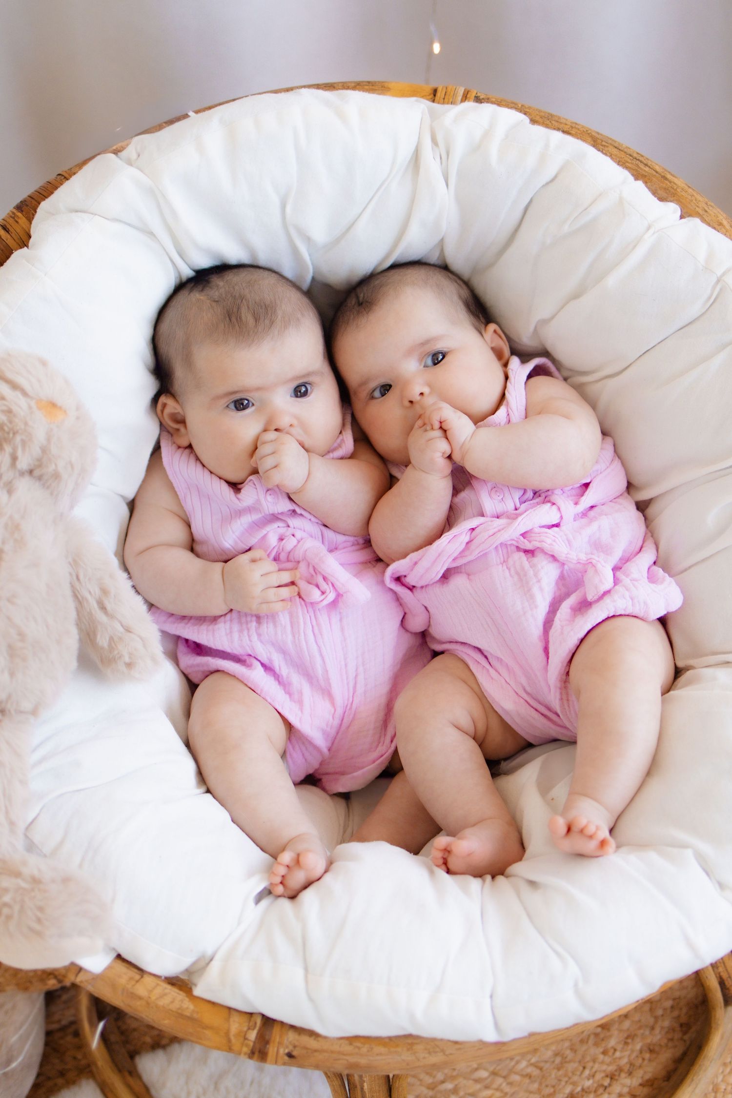 Two adorable twin baby girls in matching pink outfits lying together in a round wicker bassinet with white cushioning.