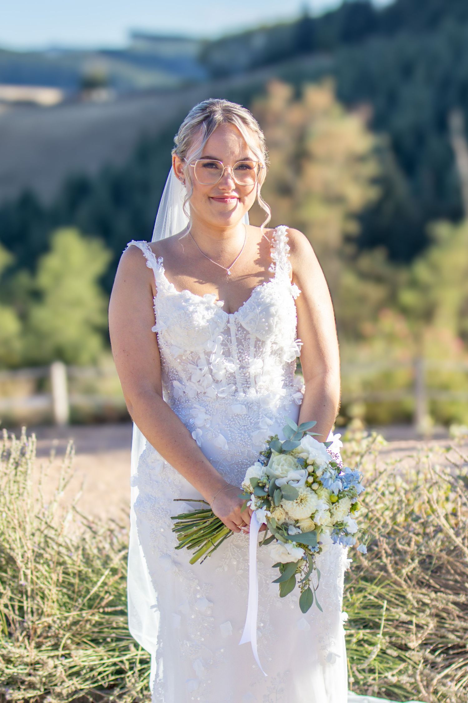 Bride in white lace gown holding bouquet of white and blue flowers, standing outdoors with rolling hills backdrop.