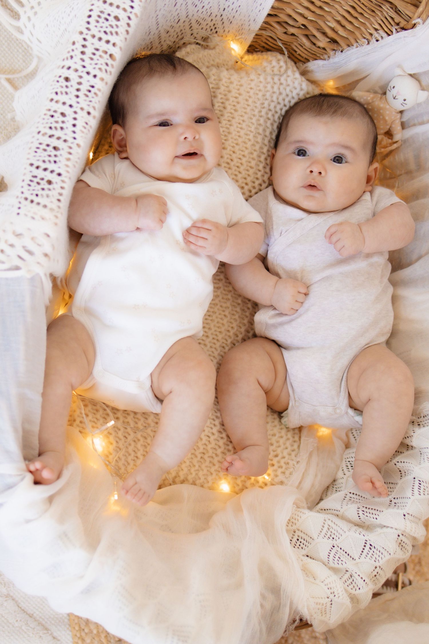 Two adorable babies in white onesies lying together in a cozy basket with warm fairy lights.