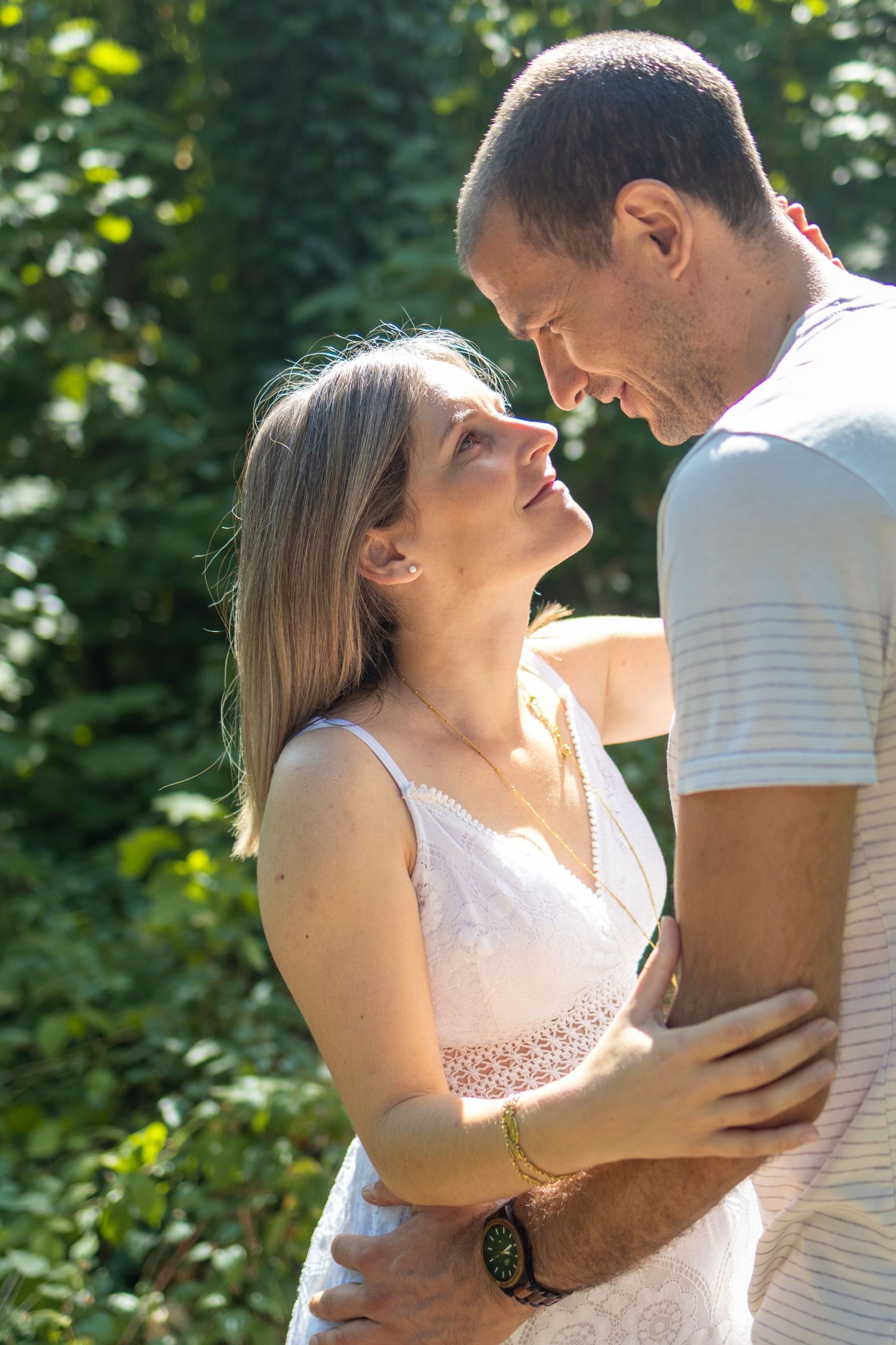 A loving couple embraces outdoors, gazing into each other's eyes surrounded by lush green foliage in warm sunlight.