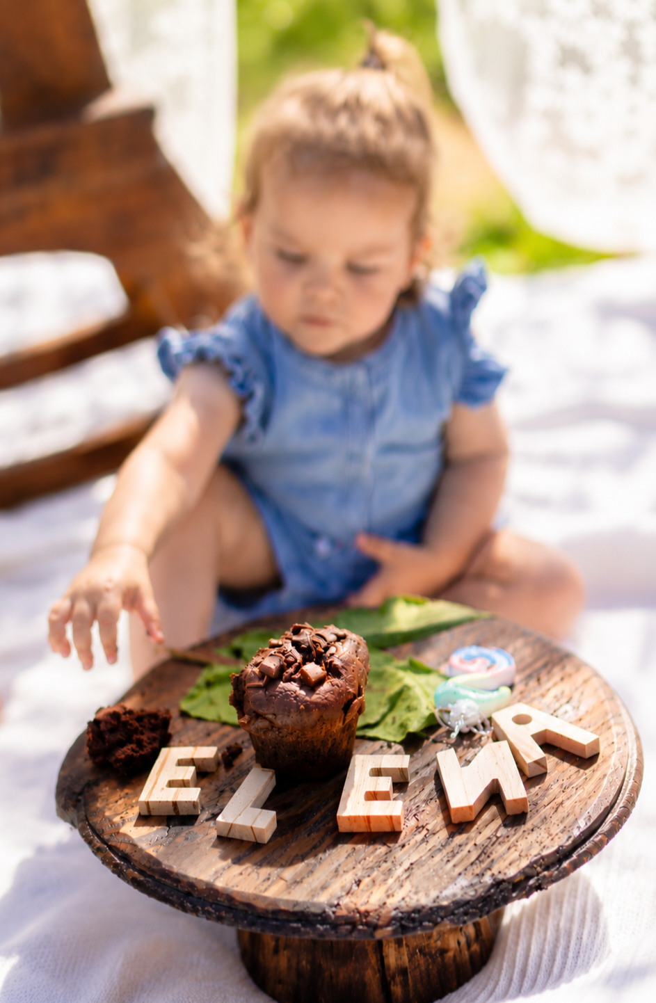 Toddler girl in denim romper reaching for chocolate muffin on rustic wooden stand with letter tiles spelling ELEMA outdoors.