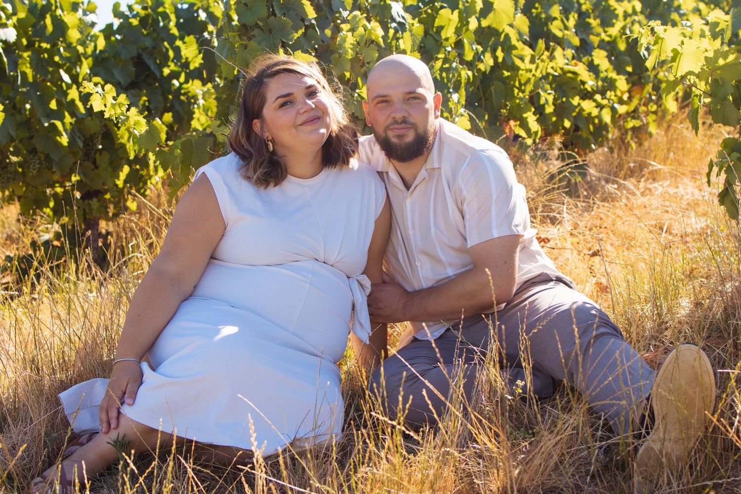 Pregnant couple sitting together in a vineyard, woman in white dress, man in beige shirt, surrounded by lush grapevines.