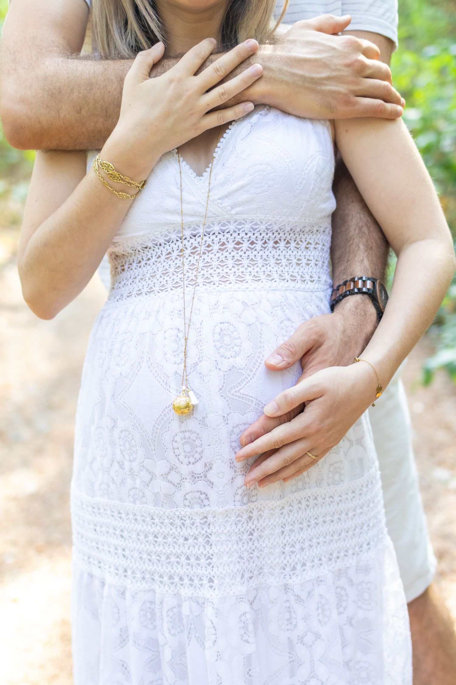 Pregnant woman in white lace dress being embraced from behind by partner, both cradling baby bump outdoors.