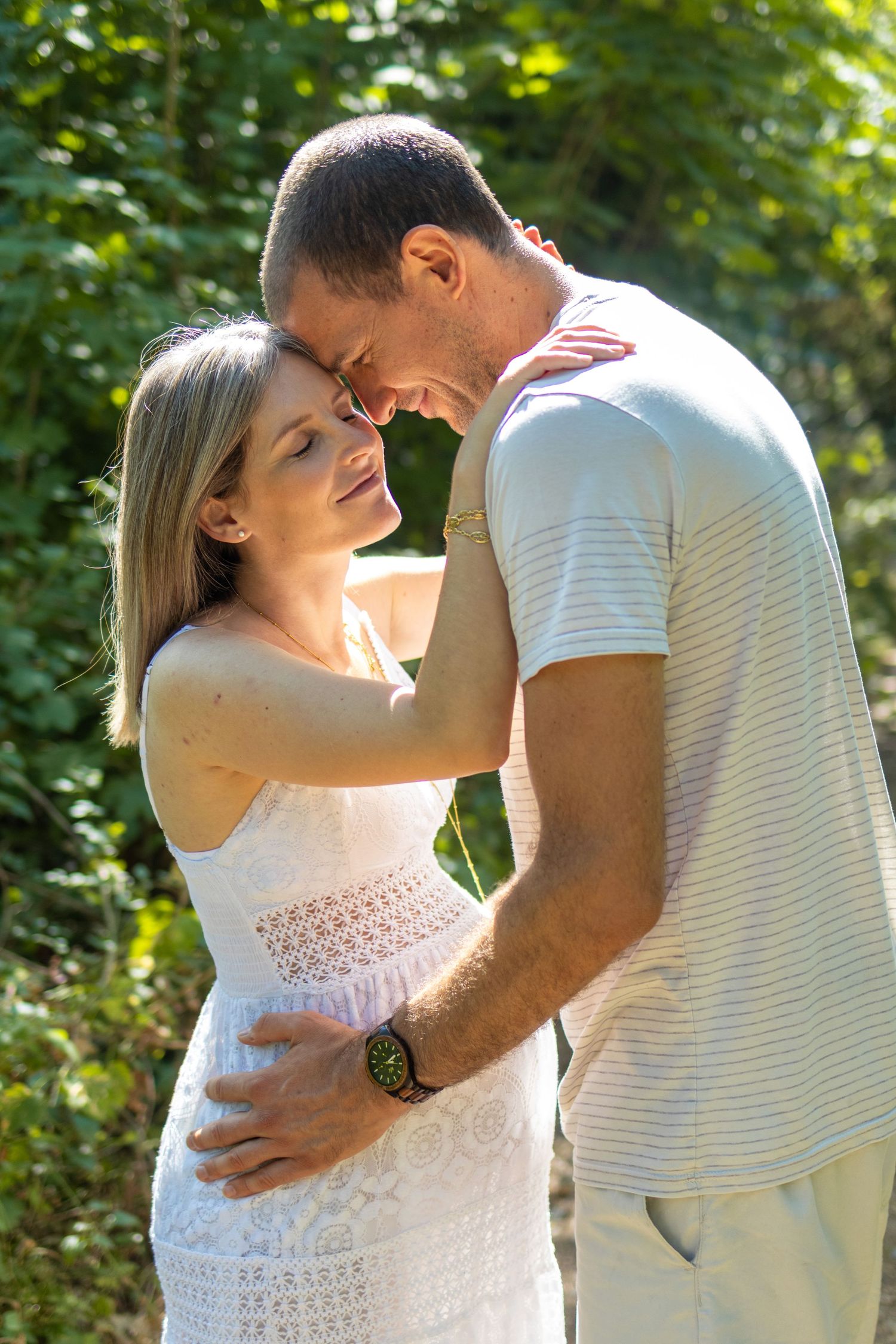 Romantic couple touching foreheads and embracing outdoors, surrounded by lush green trees on a sunny day.