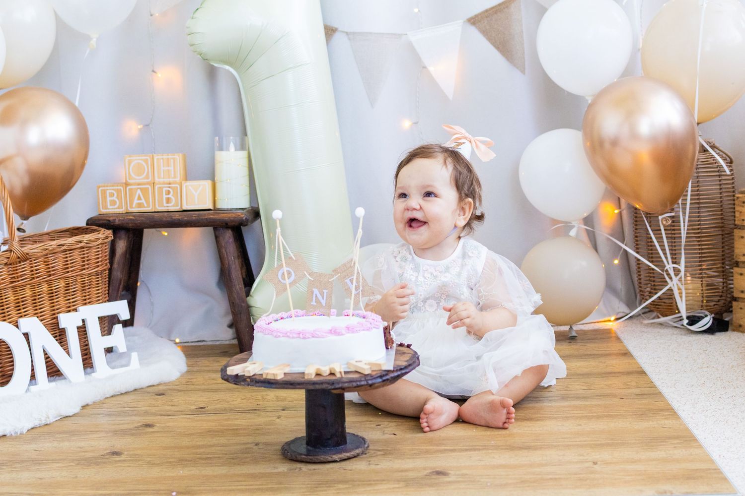 Baby girl in white dress sits beside a pink first birthday cake, surrounded by balloons and ONE sign decorations.