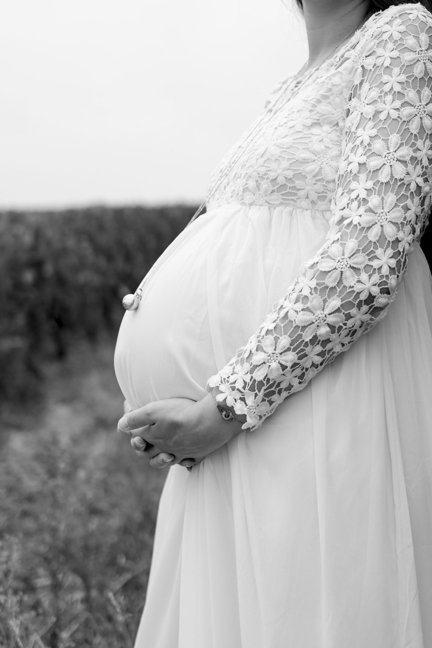 Black and white photo of a pregnant woman in a white lace dress cradling her baby bump outdoors.