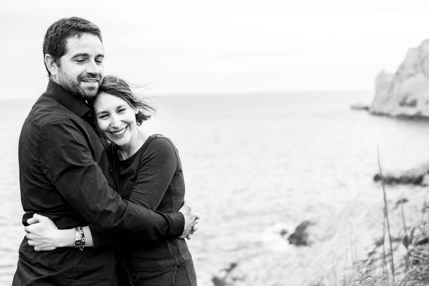 Black and white photo of a happy couple embracing and smiling by the ocean shore.