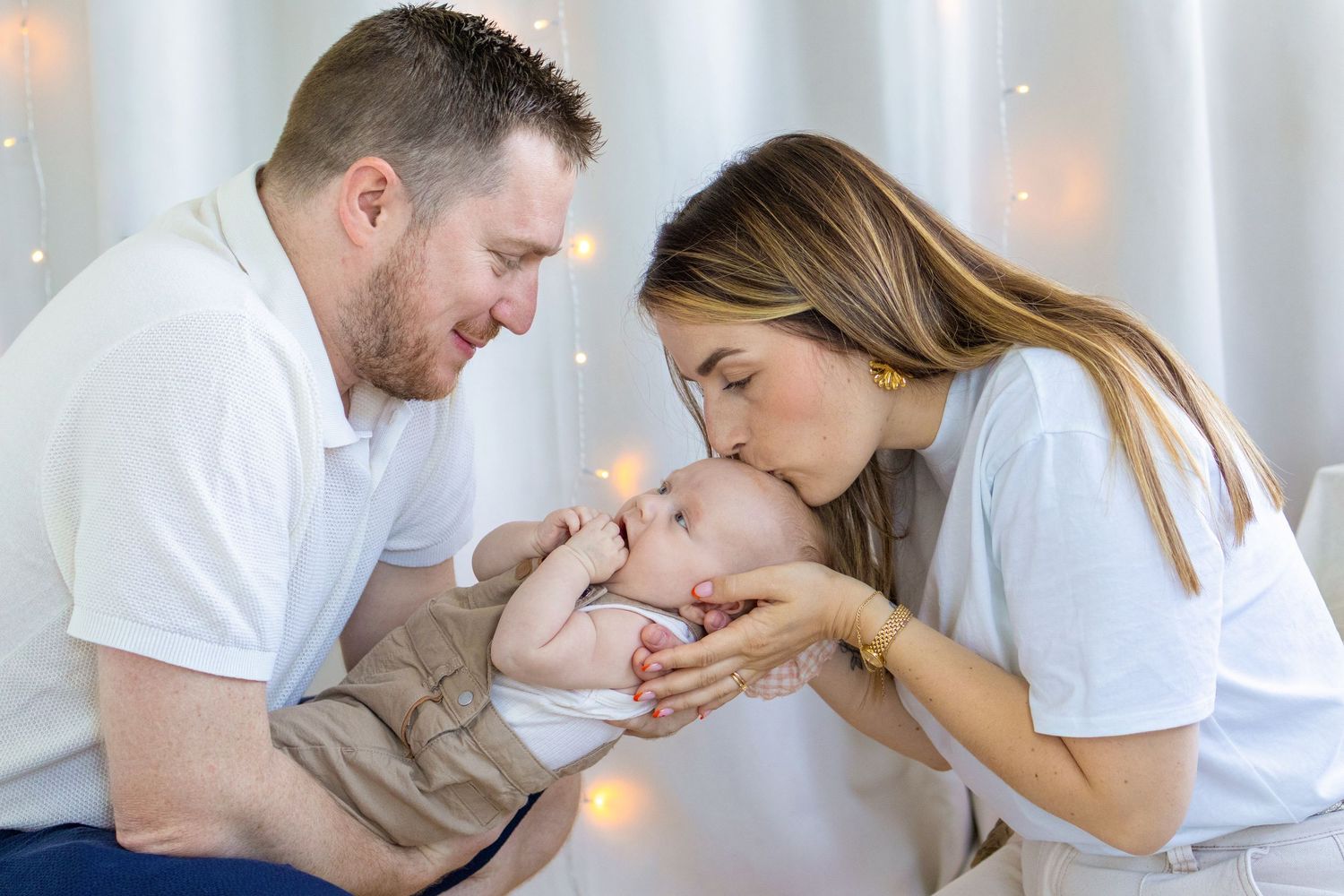 Happy parents in white shirts kissing and bonding with their newborn baby amid soft bokeh lights.