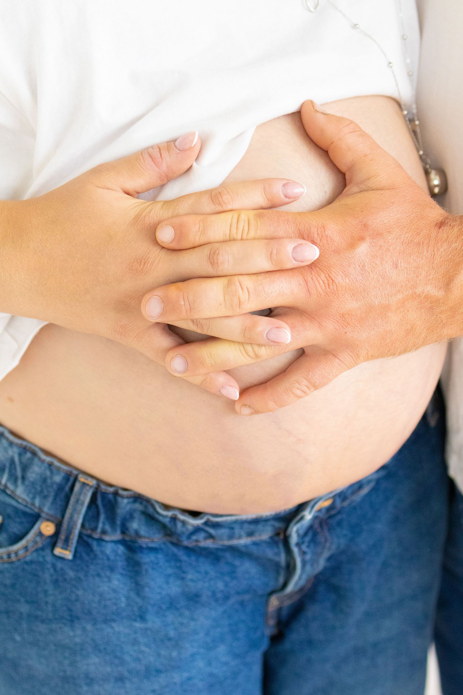 Two pairs of hands gently cradling a pregnant woman's bare belly, wearing blue jeans and white top.