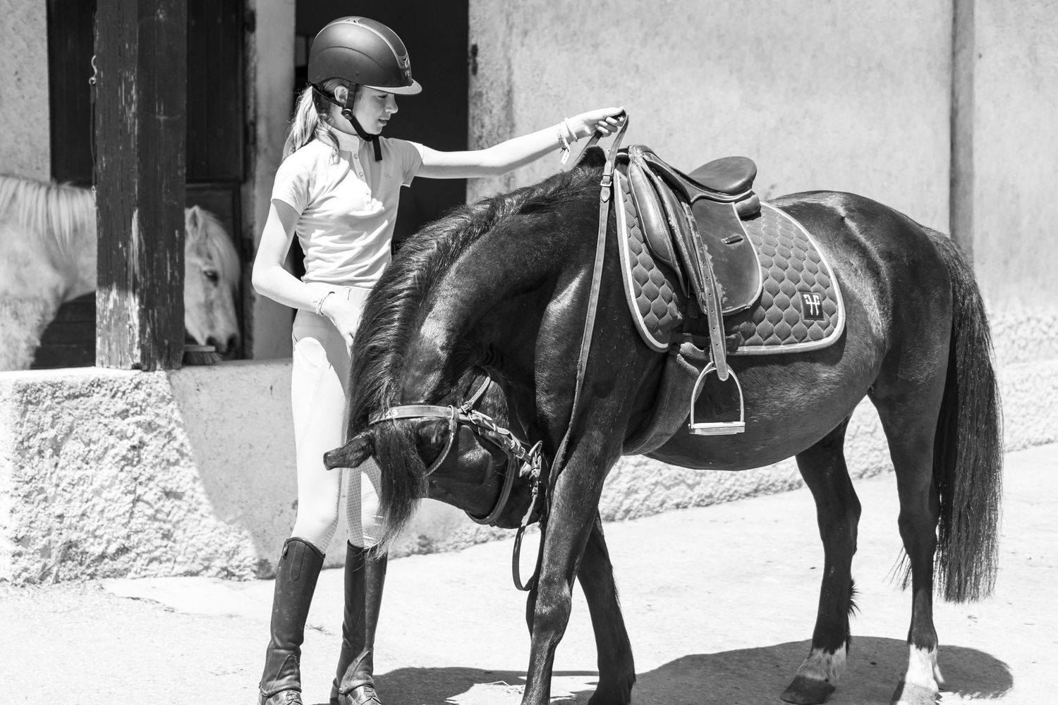 Young female equestrian in riding gear petting a dark horse outdoors in black and white photo.